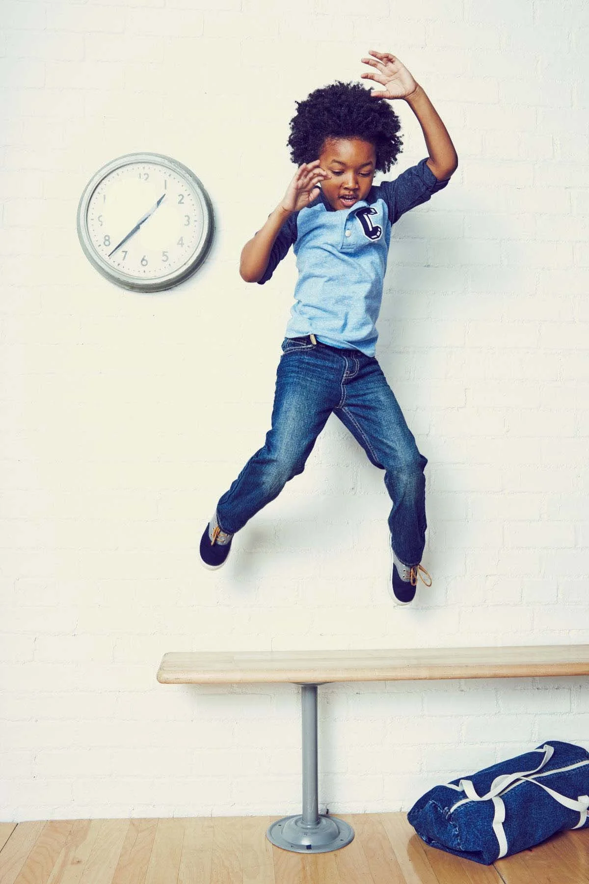 A young boy with curly hair jumping above a balance beam in a room with a white brick wall, a clock showing around 1:08, and a blue and white bag on the floor.