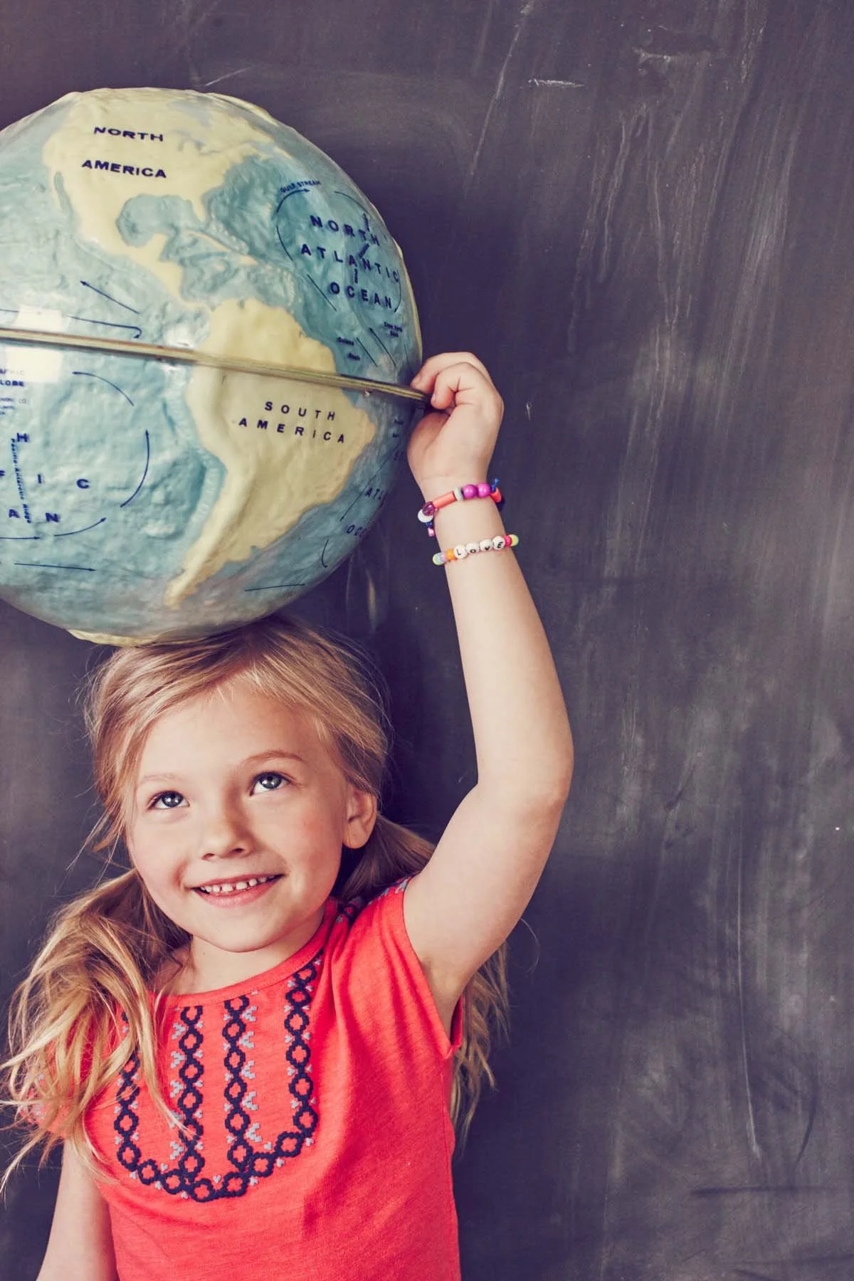 A young girl with blonde hair smiling while holding a globe of the Earth above her head, with her eyes looking upward. She is wearing an orange shirt with navy embroidery and colorful beaded bracelets on her wrist. The background is a dark, textured 
