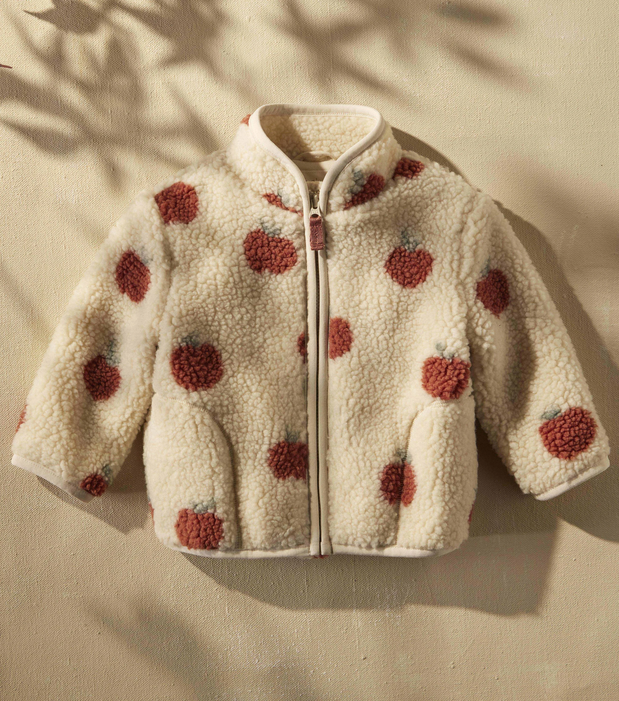 A fluffy, cream-colored fleece jacket with red and gray polka dots, zipped up and displayed against a beige fabric background.