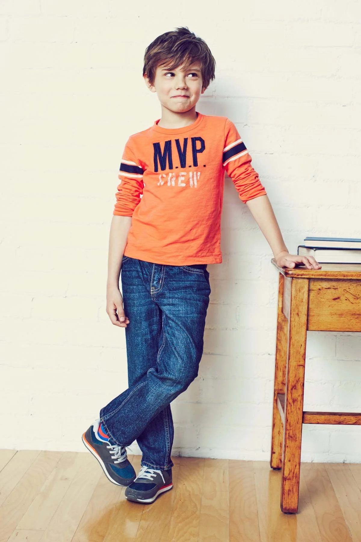 A young boy with tousled brown hair wearing a bright orange long-sleeve shirt that says "M.V.P. Crew" in navy and white, blue jeans, and gray sneakers with blue and orange accents, leaning against a white brick wall with a wooden table nearby, on whi