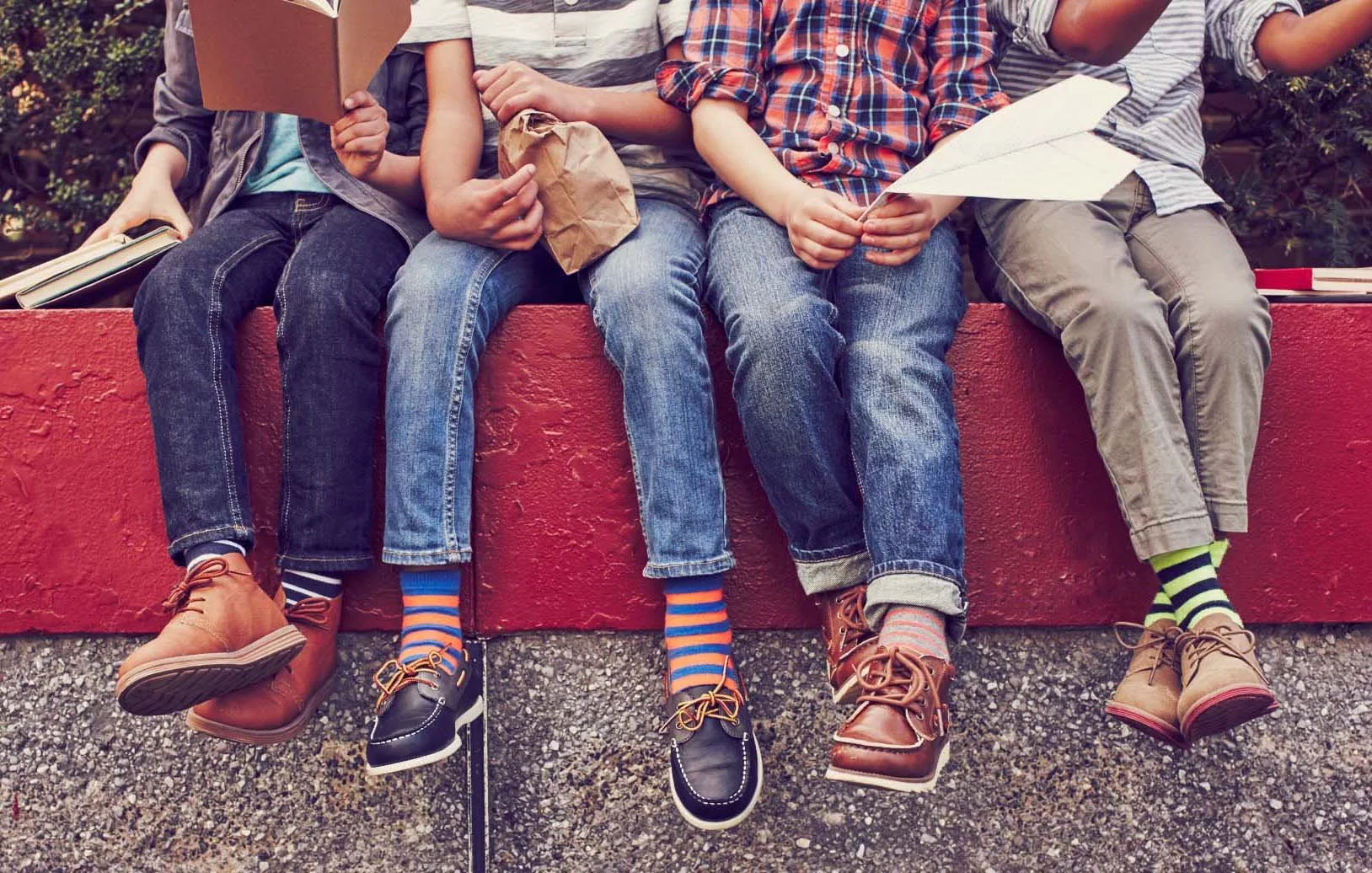 Four children sitting on a red ledge reading books and holding papers, wearing colorful striped socks and boots, with green bushes in the background.