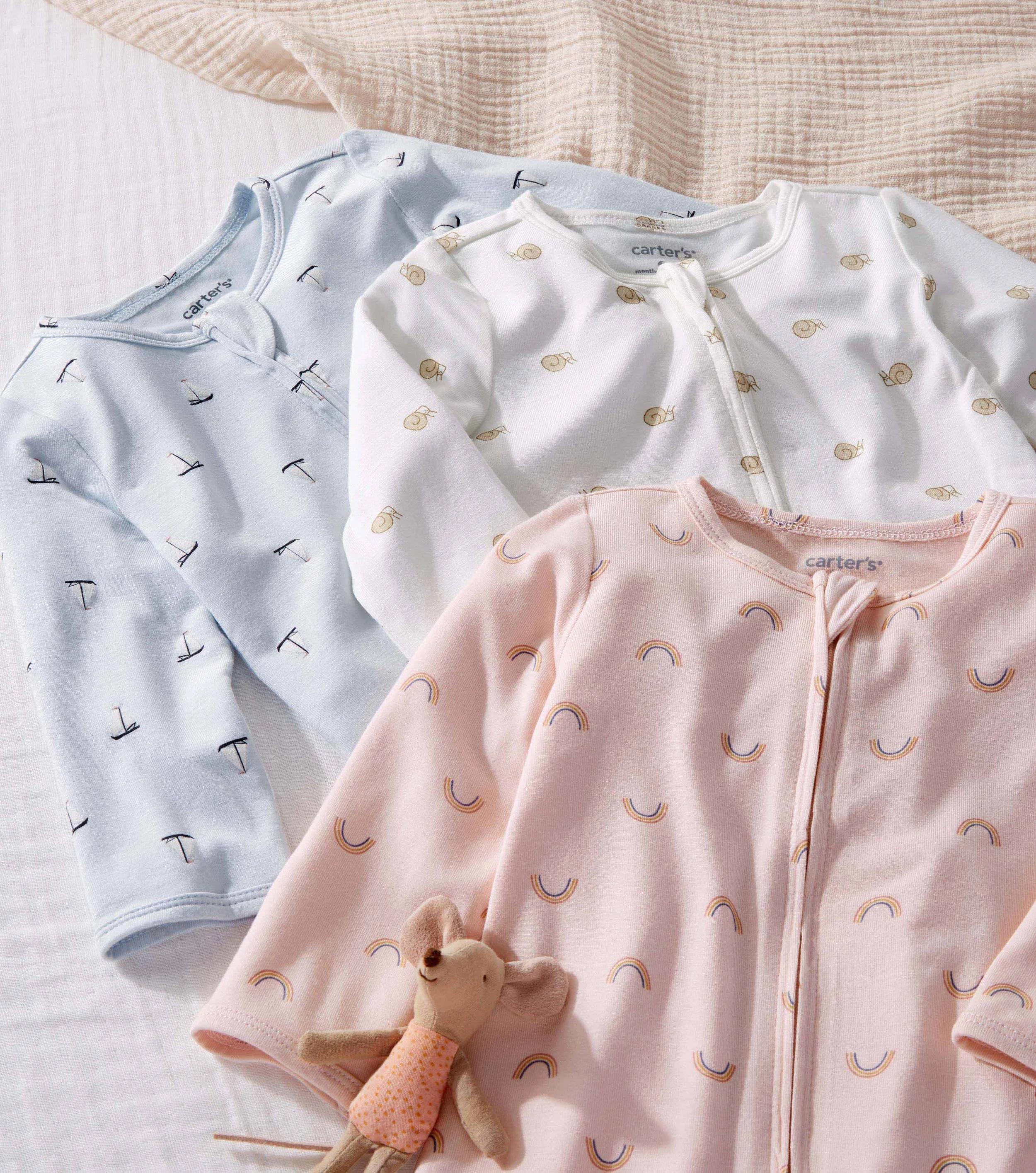 Purely Soft pajamas in pastel-colored with patterns of rainbows and snails. The pajamas have front zippers and cuffs at the sleeves and ankles. A small stuffed bear toy is placed at the bottom of the pajamas.
