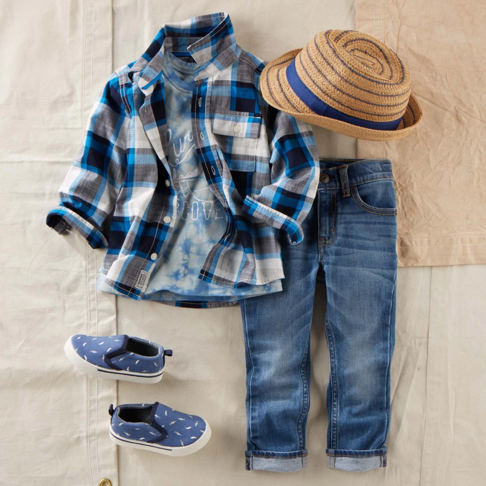 A folded plaid shirt, a collared T-shirt, a straw hat with a blue ribbon, a pair of jeans, blue sneakers with white soles and laces, and a pair of black sneakers with white soles on a light colored fabric surface.