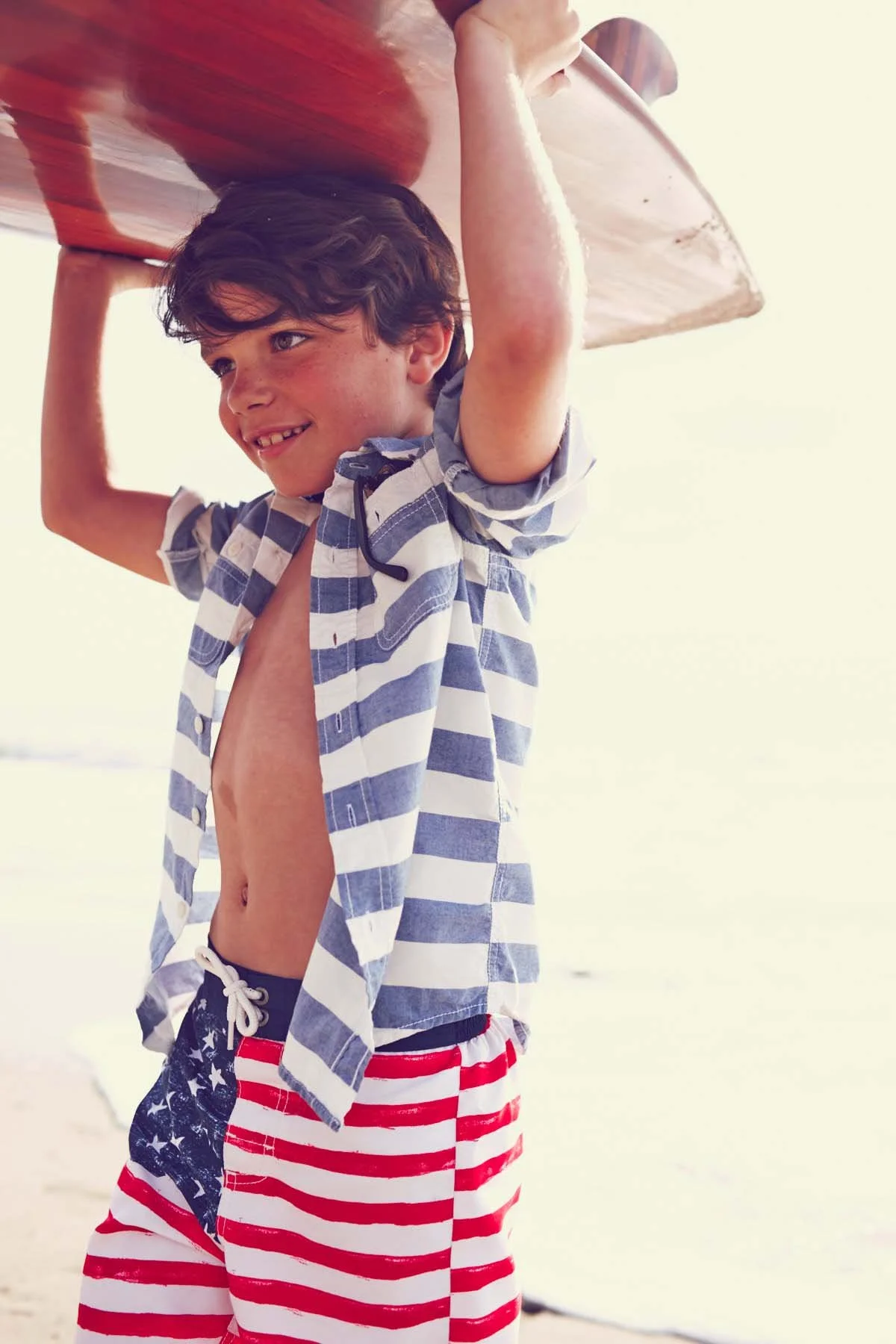 A young boy smiling, holding a surfboard above his head at the beach, wearing a blue and white striped shirt and American flag shorts.