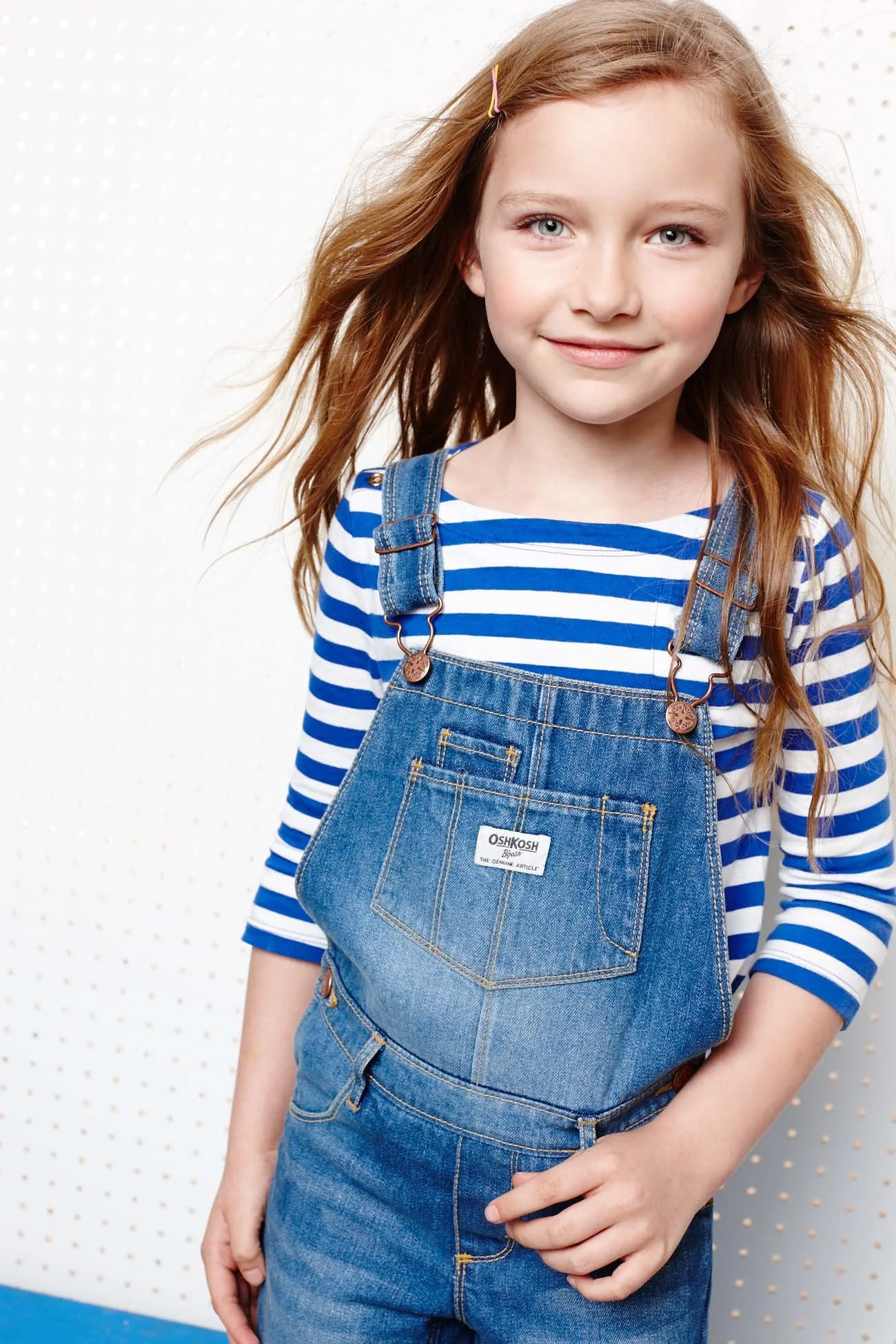 A young girl with red hair wearing a blue and white striped shirt and denim overalls.
