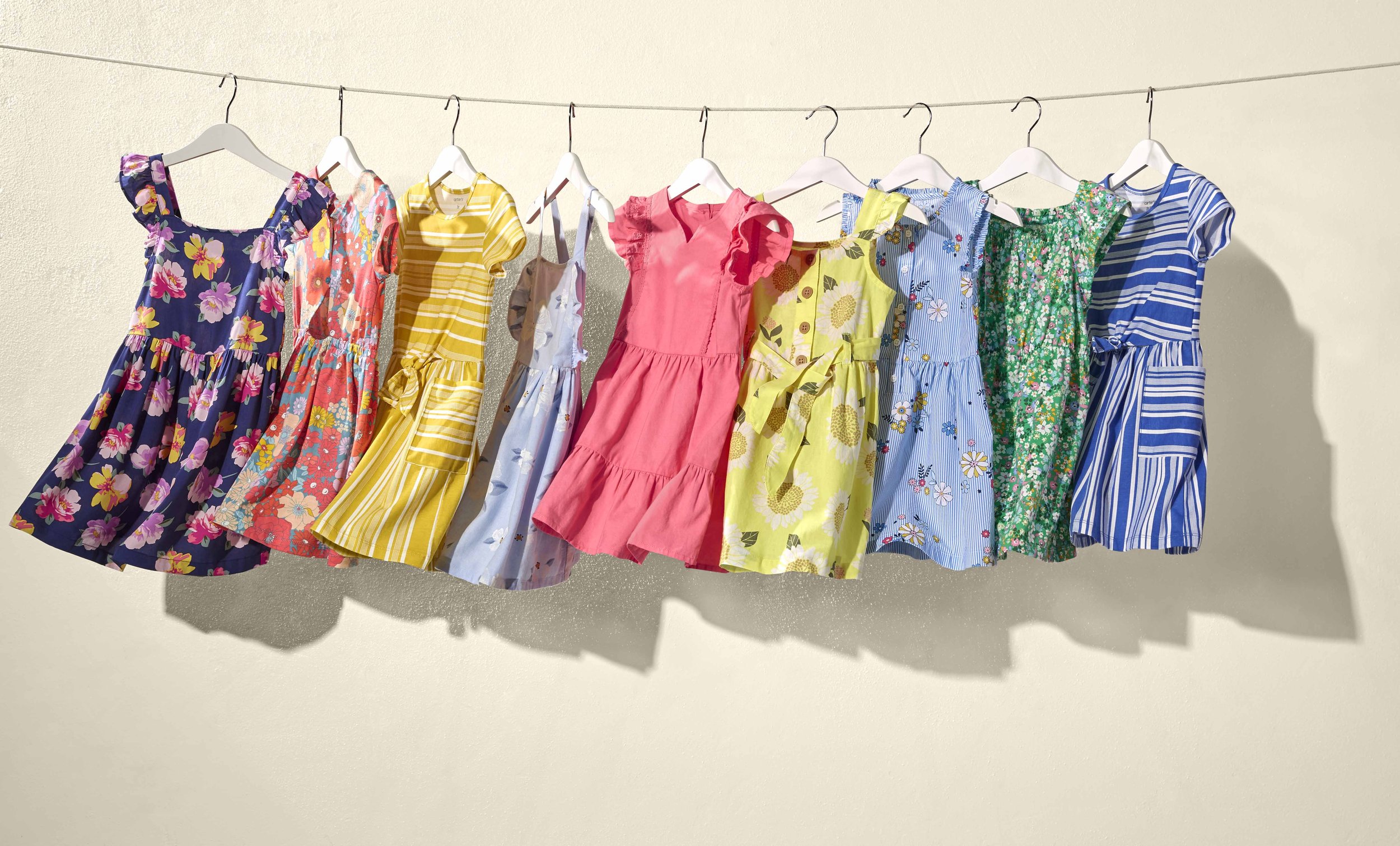 A row of colorful dresses with floral and striped patterns hanging on a wire against a beige wall.
