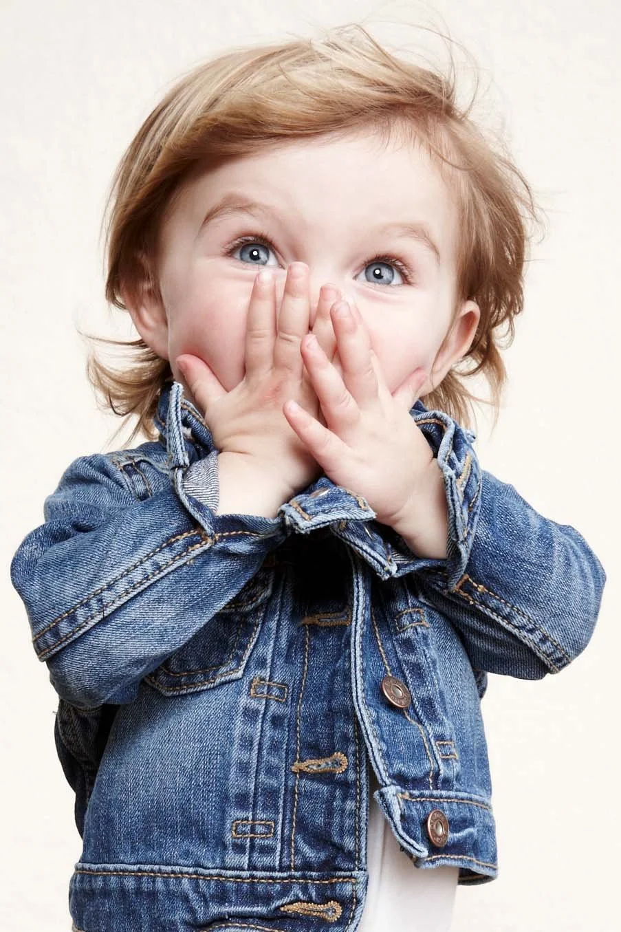 A young child with light brown hair and blue eyes covering their mouth with hands, wearing a denim jacket.