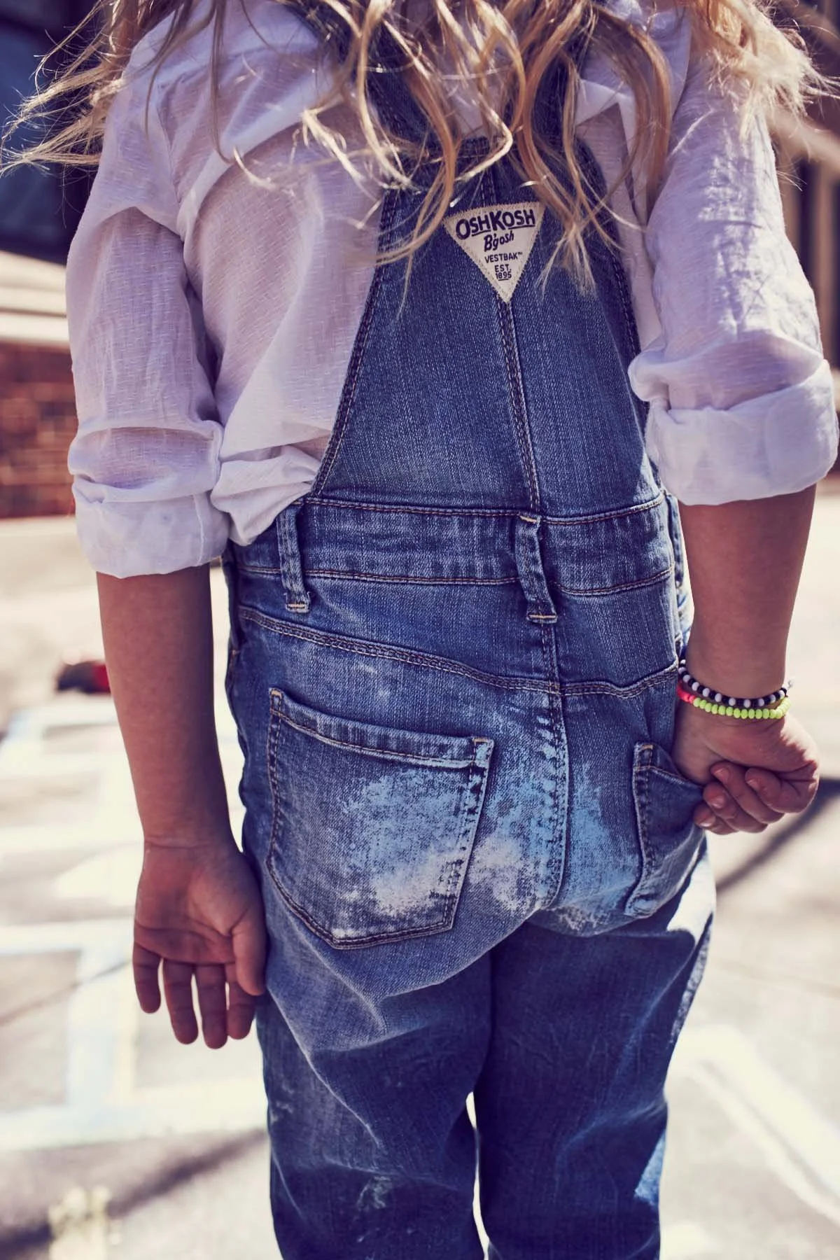 Close-up of person wearing a white, long-sleeve shirt rolled up at the sleeves, denim overalls, and beaded bracelets, standing outdoors on a sunny day.