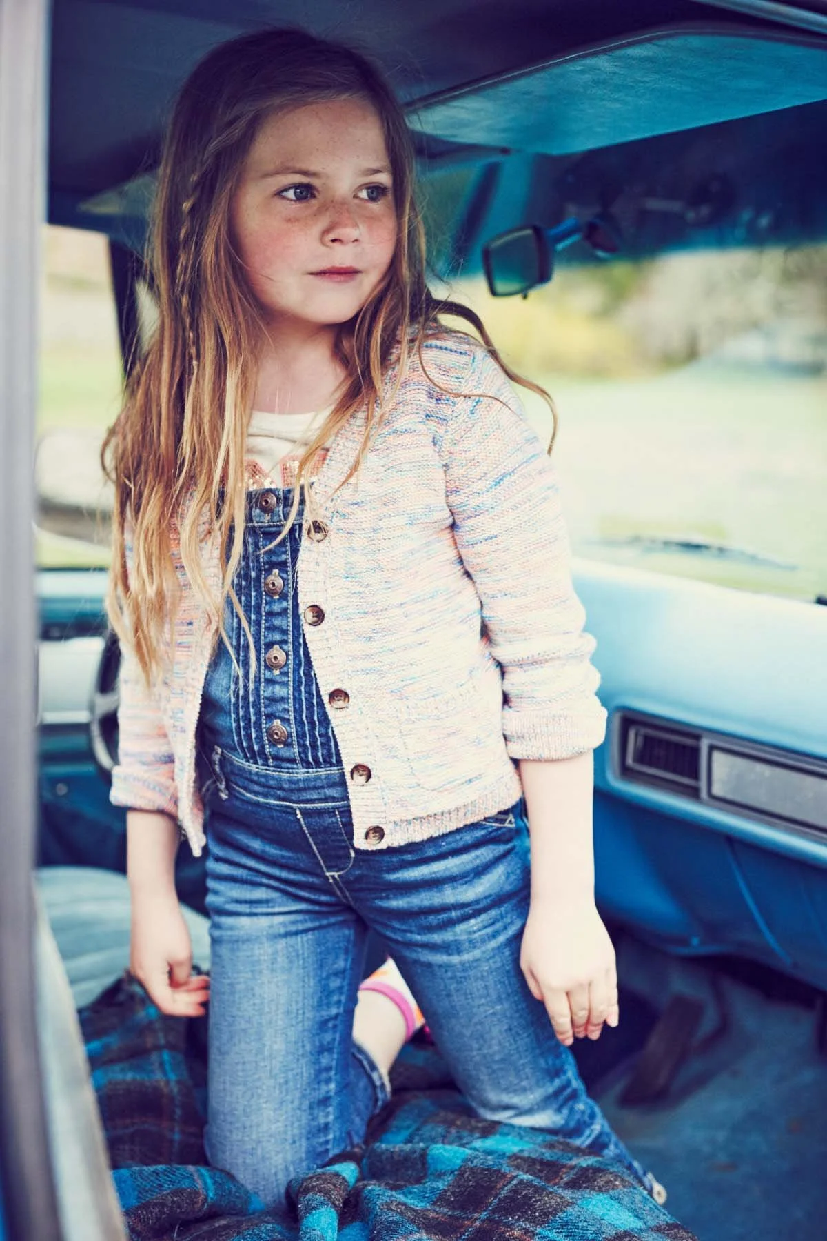 A young girl with long, wavy red hair and freckles stands partially inside a vehicle, looking to her left. She wears a colorful, striped cardigan over denim overalls and jeans, with someone lying down underneath a blanket in the foreground.