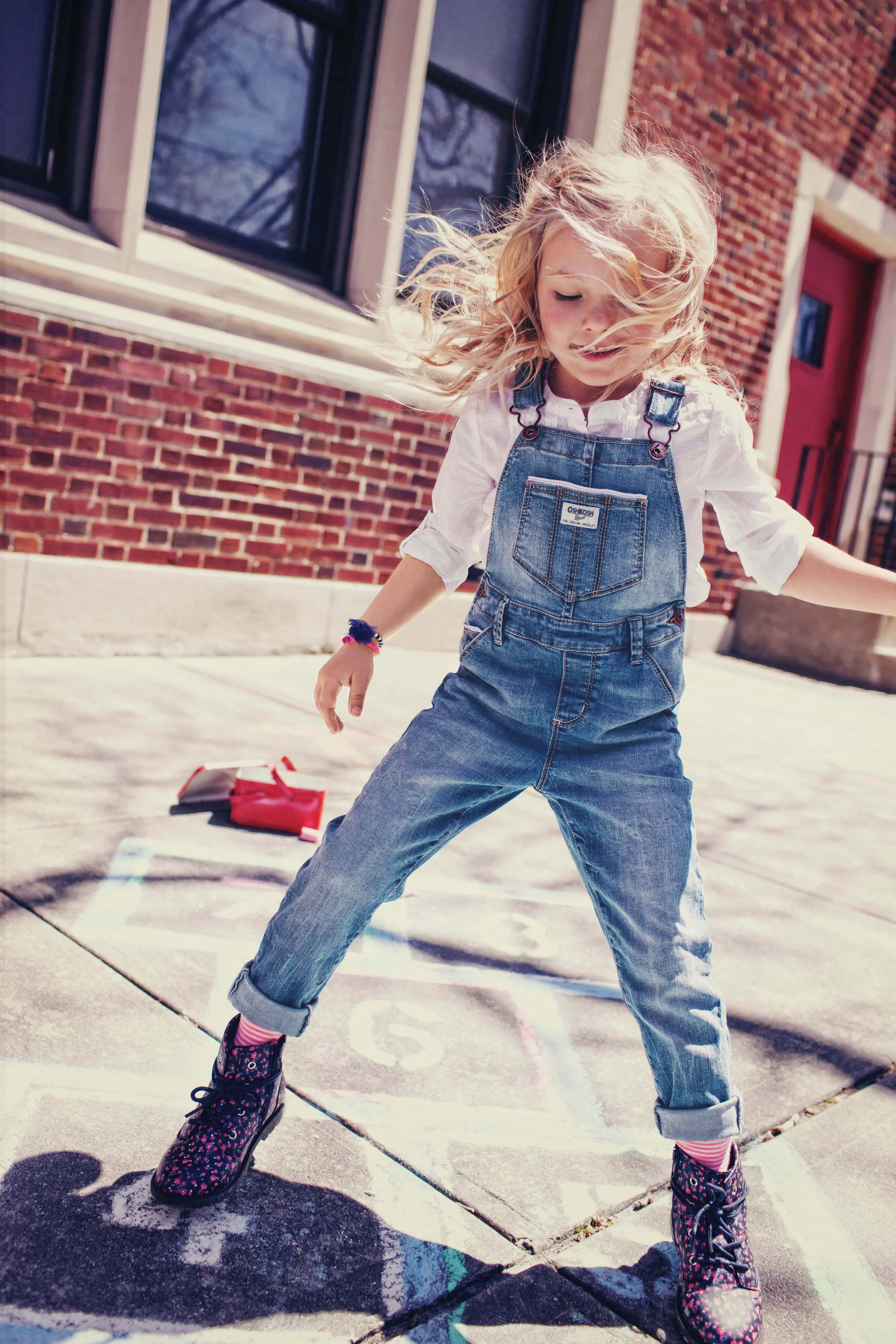 A young girl with blonde curly hair is roller skating outdoors on a concrete surface marked with hopscotch. She is wearing denim overalls, a white shirt, and patterned roller skates, with a playful expression on her face.