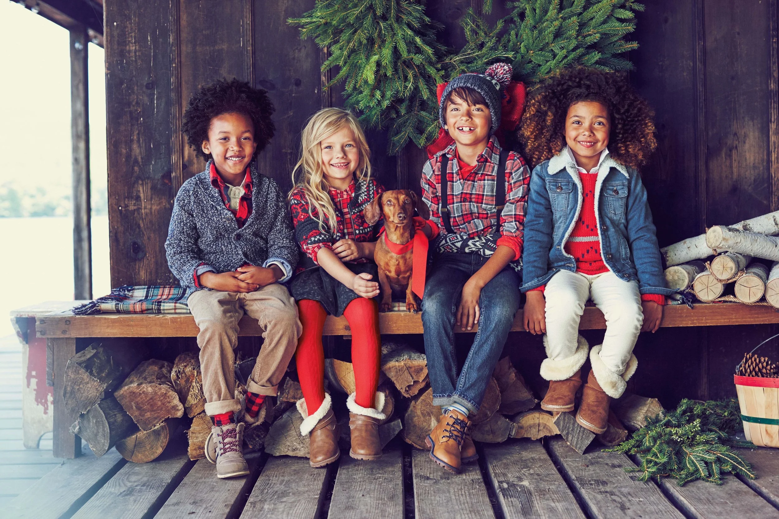 Five children sitting on a wooden bench in front of Christmas trees, dressed in winter clothing with festive, holiday-themed attire, and a small brown dog in the center.