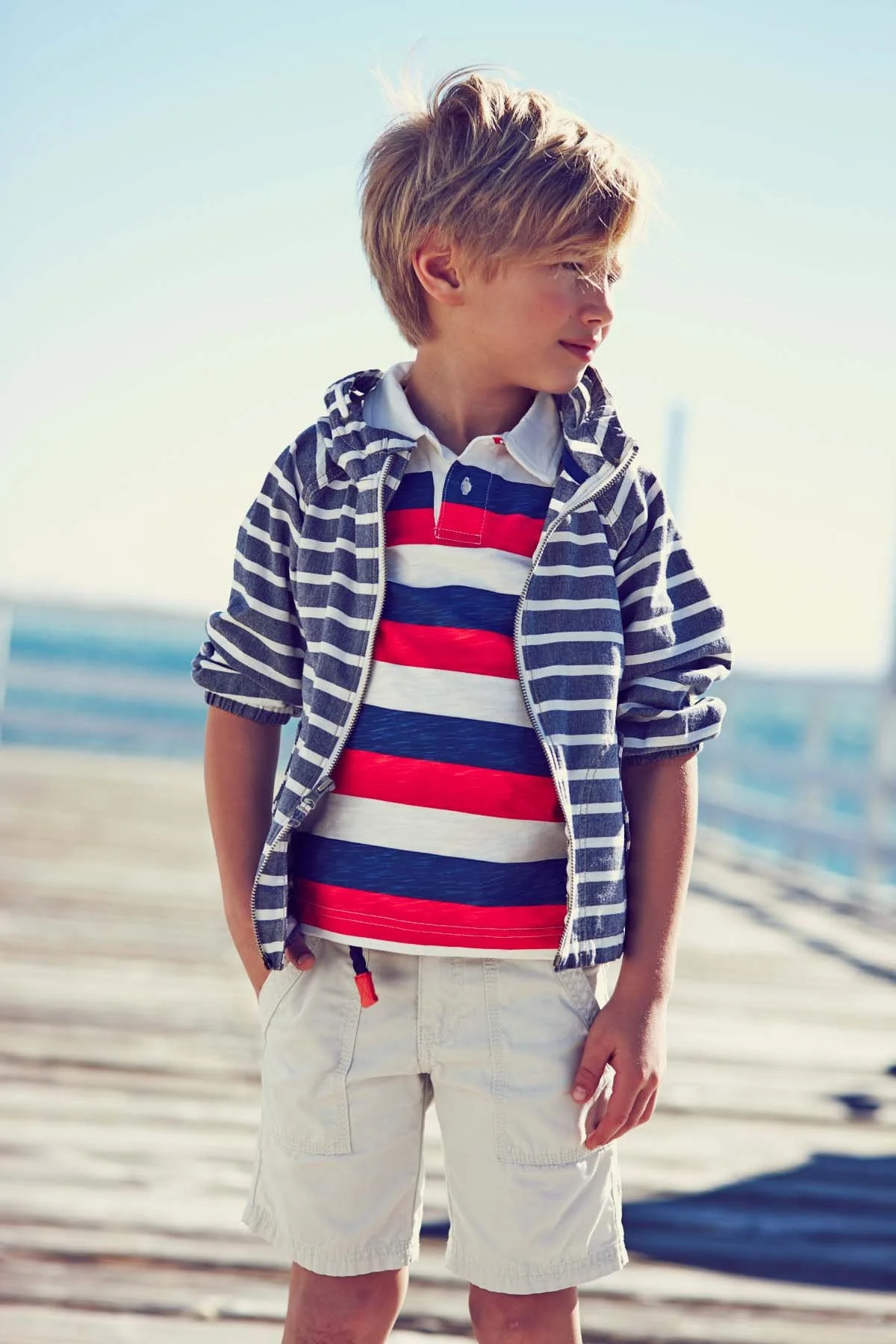 A young boy standing on a wooden dock at the beach, wearing a striped polo shirt, a zip-up hoodie with horizontal stripes, and beige shorts, with the ocean in the background.