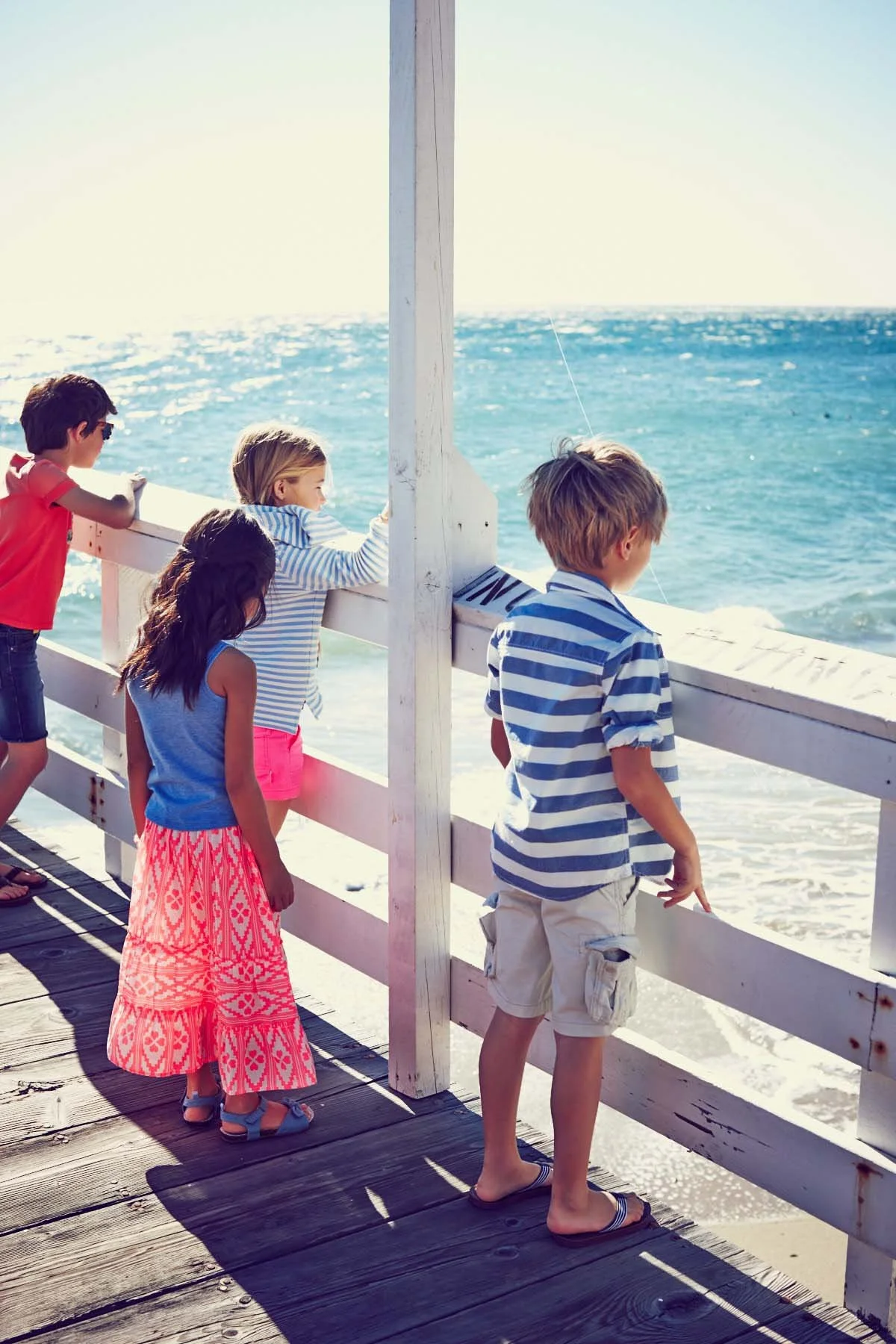 Four children looking out over the ocean from a wooden pier, bathed in sunlight, some wearing striped clothing and swimwear, with waves crashing below.