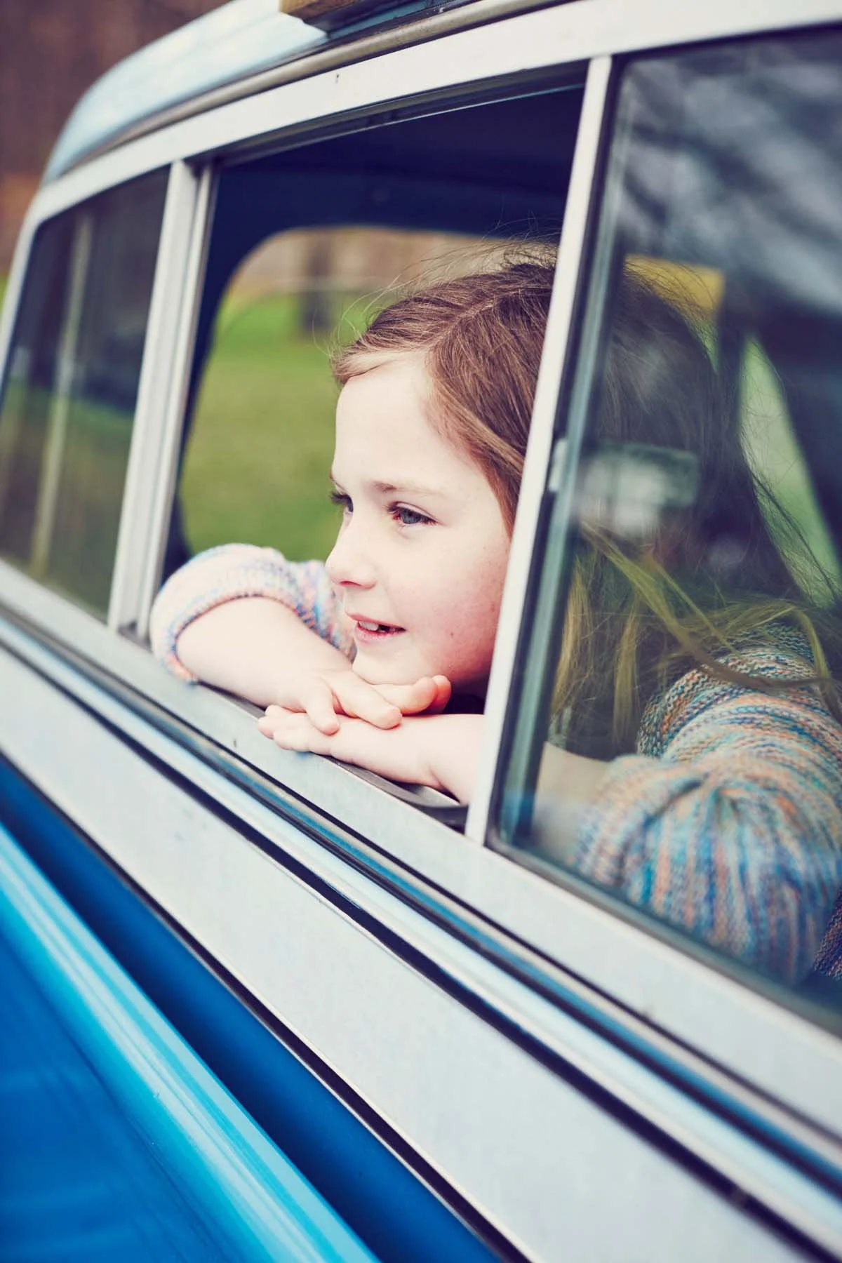 A young girl with brown hair and blue eyes leaning on the window sill of a blue and white vintage car, looking outside with a peaceful expression.