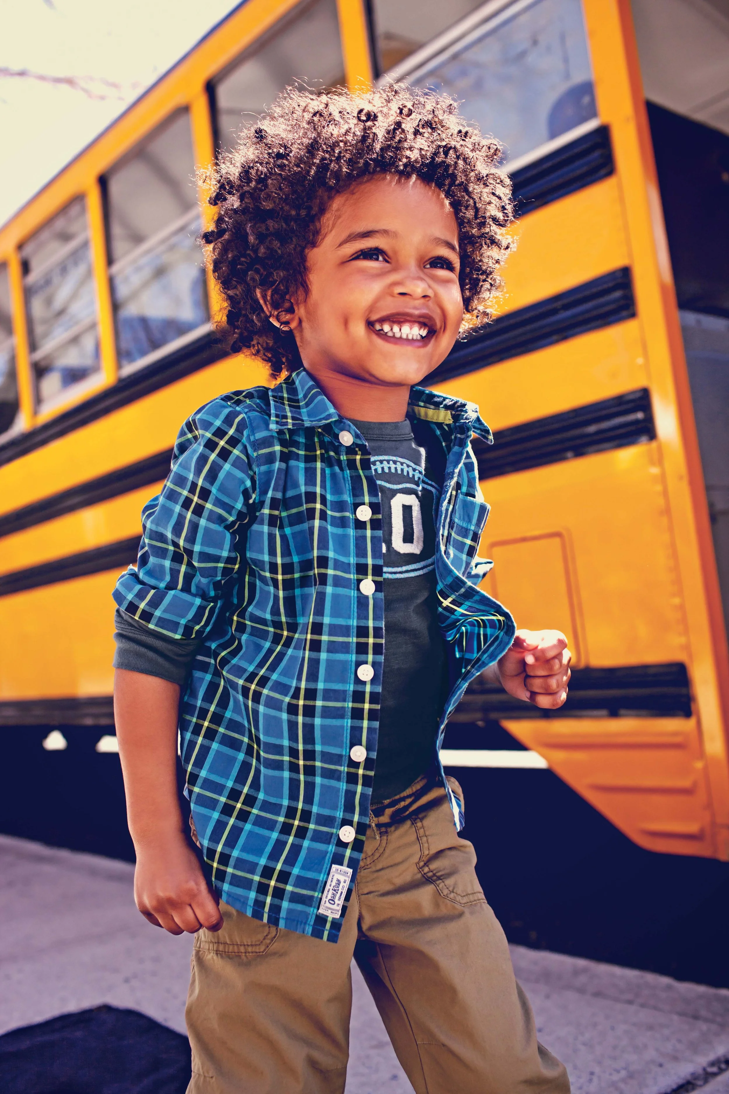 A young boy with curly hair smiling and running near a yellow school bus, wearing a blue plaid shirt and khaki pants.
