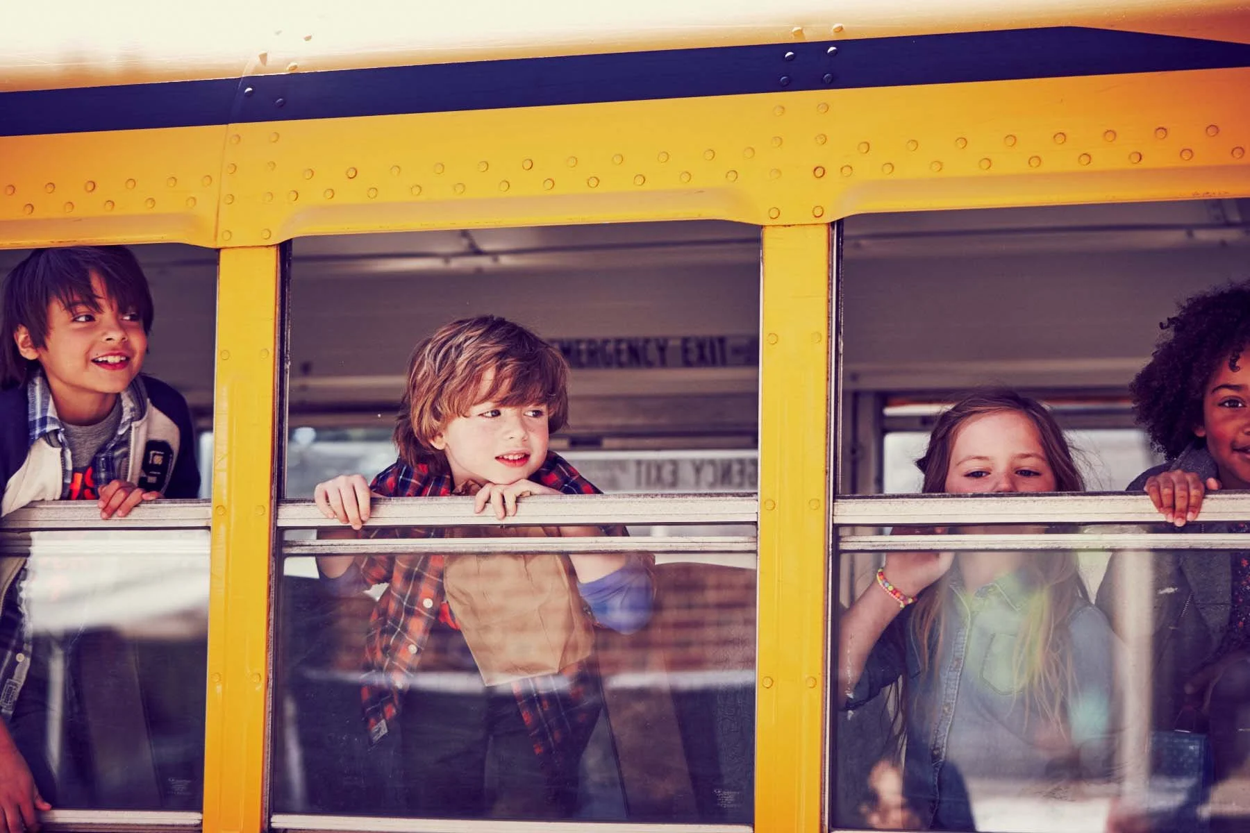 Four children leaning out of a school bus window, looking outside with curiosity and smiles.