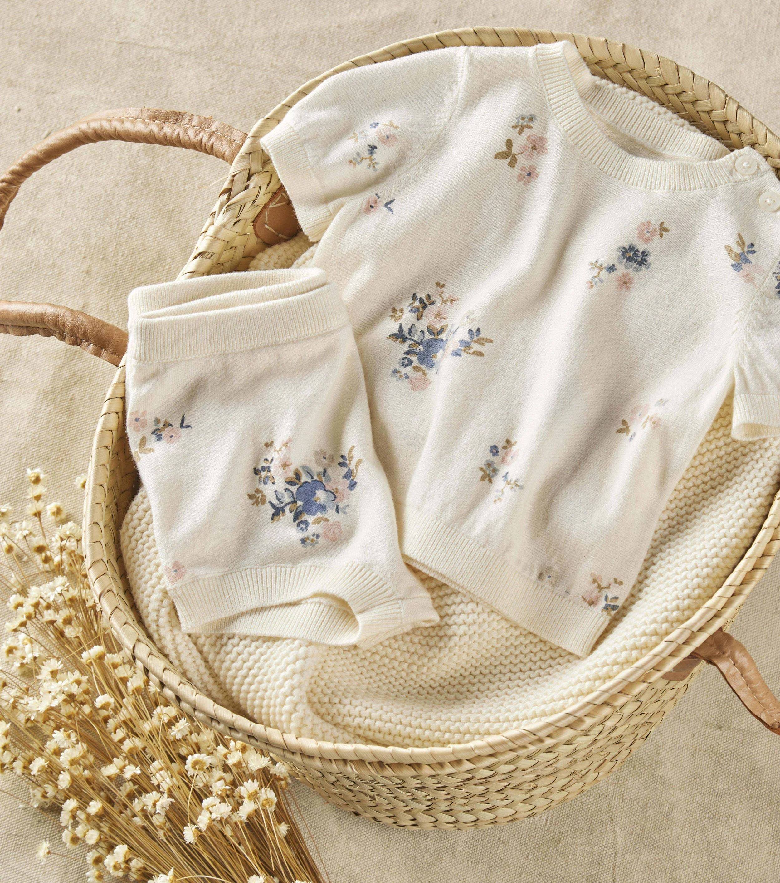 A soft white baby sweater with a floral pattern, folded and placed in a round wicker basket on a beige textured surface, with a bunch of dried small white flowers nearby.