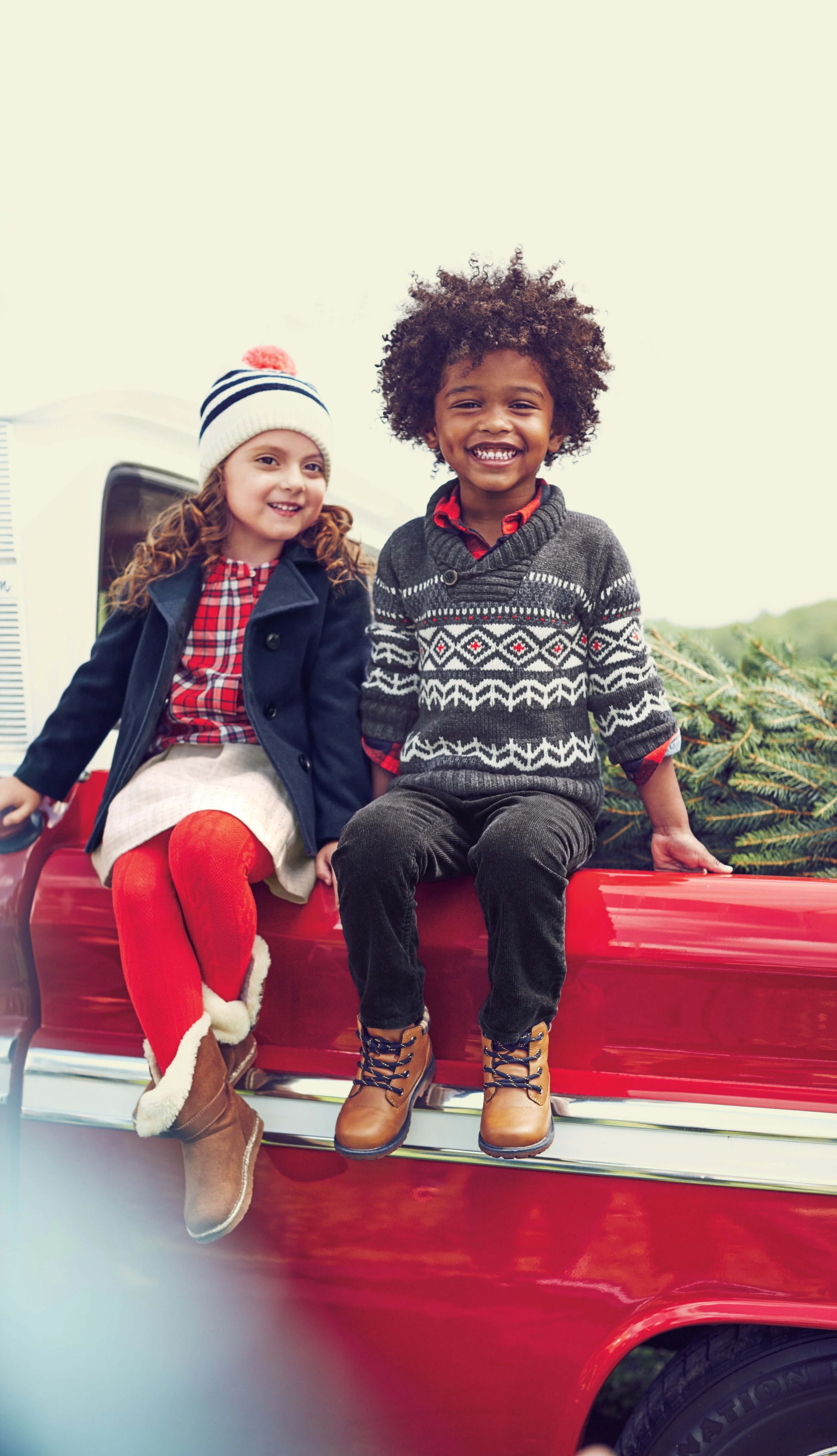 Two children, a girl and a boy, sitting on the edge of a red vehicle, smiling. The girl has curly red hair, a beanie, and a navy coat. The boy has curly hair, a patterned sweater, and tan boots. There are Christmas tree branches in the background.