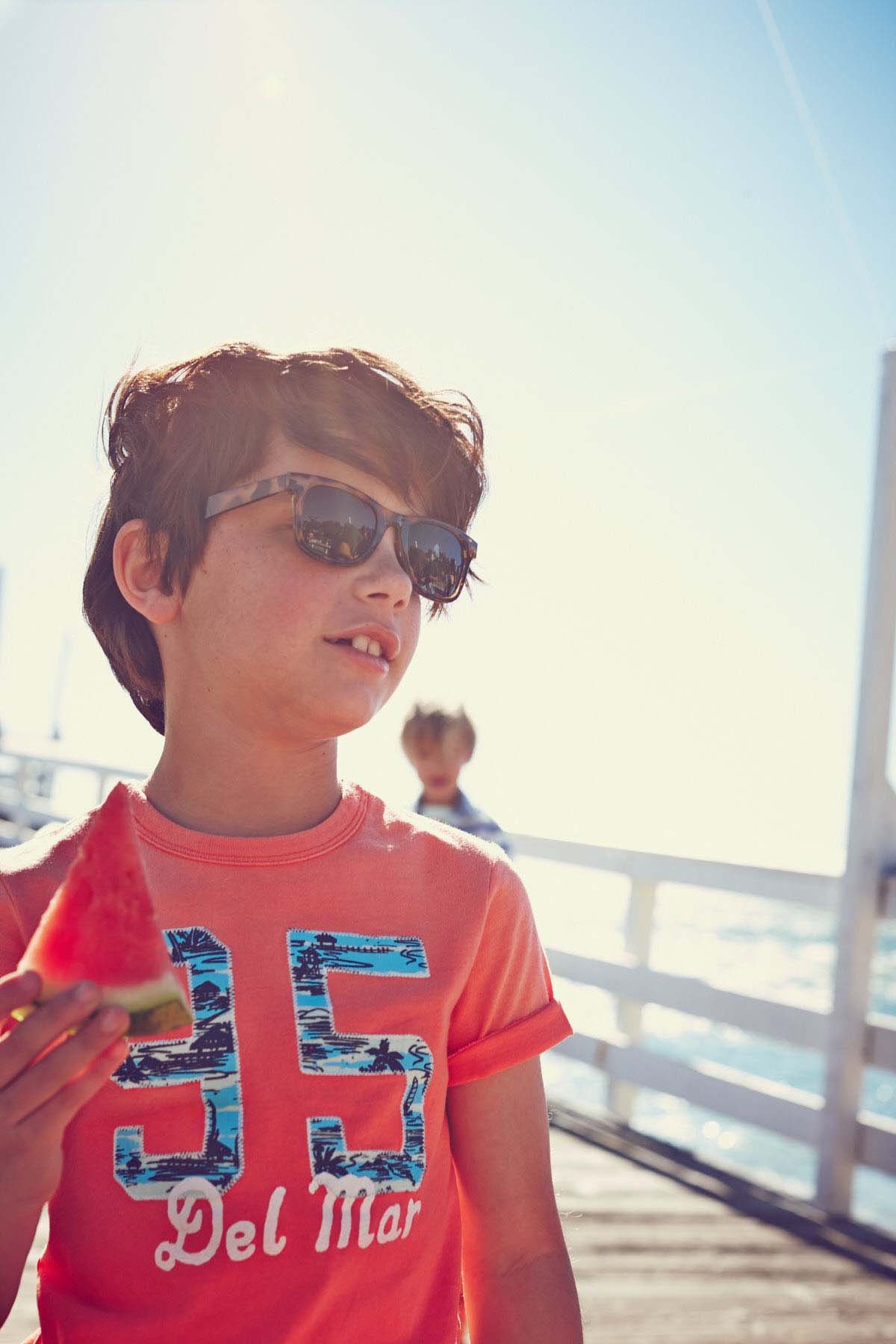 A young boy with brown hair wearing sunglasses and an orange T-shirt holding a slice of watermelon on a sunny day, with another child in the background near a white fence by the water.