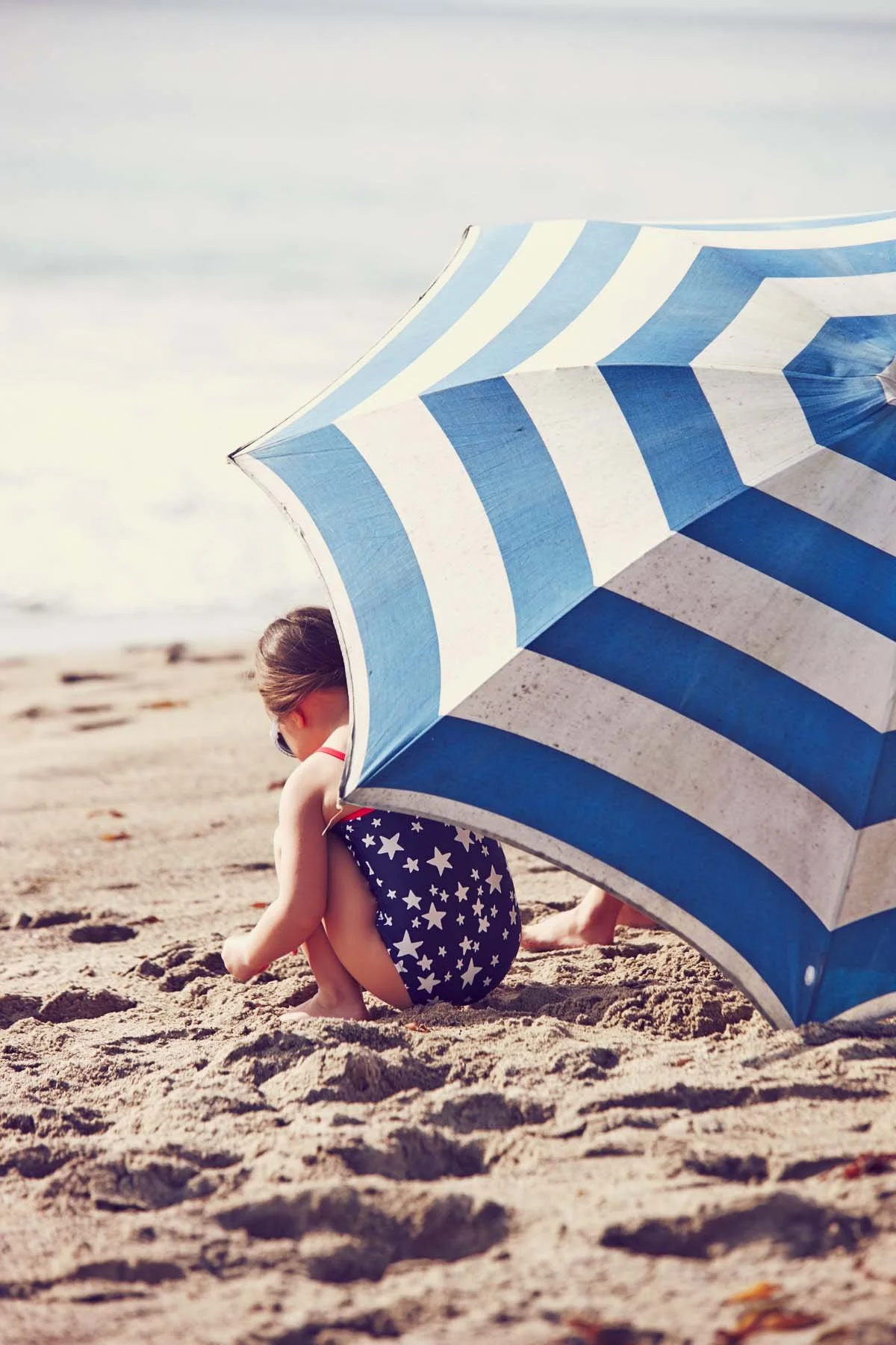 A young girl with a star-patterned swimsuit sitting on a sandy beach near a blue and white striped beach umbrella, looking down at the sand.
