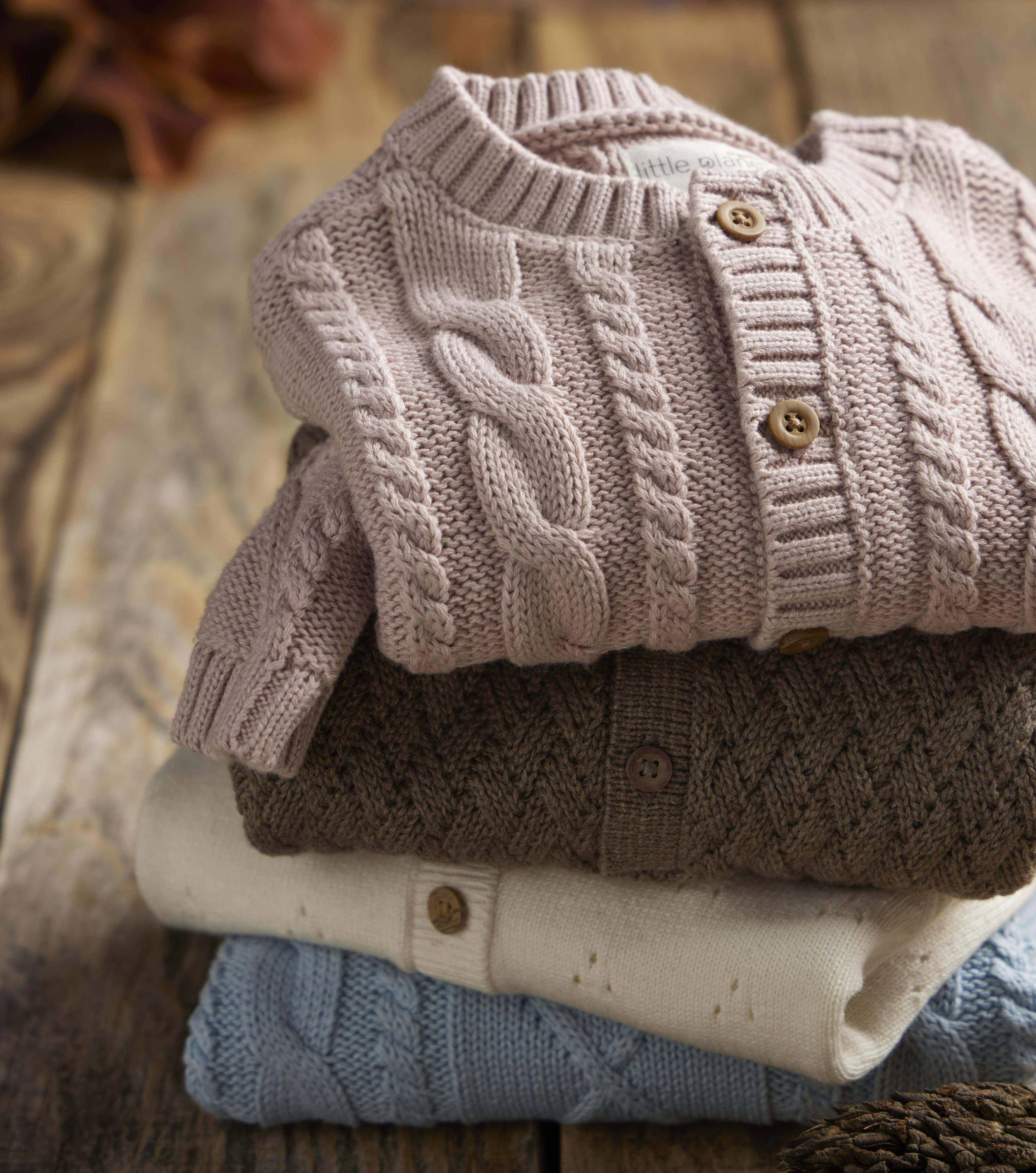 Three folded knitted sweaters in beige, white, and light blue colors stacked on a wooden surface.