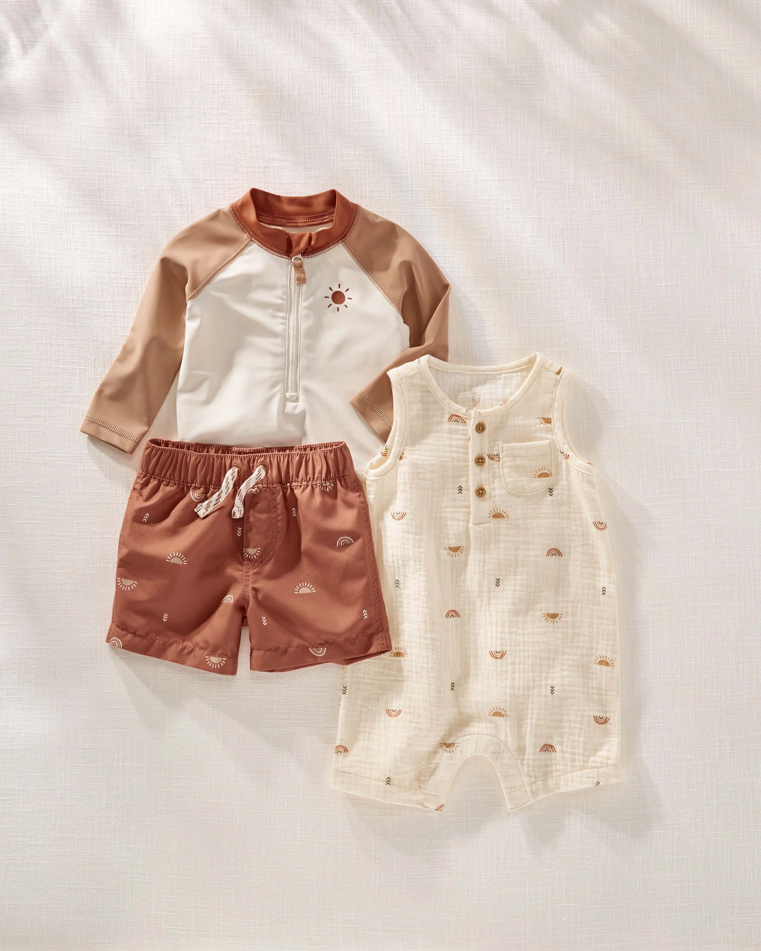 A set of children's clothing on a light-colored textured background, including a long-sleeve shirt, a sleeveless dress with sun and rainbow patterns, and a pair of shorts.