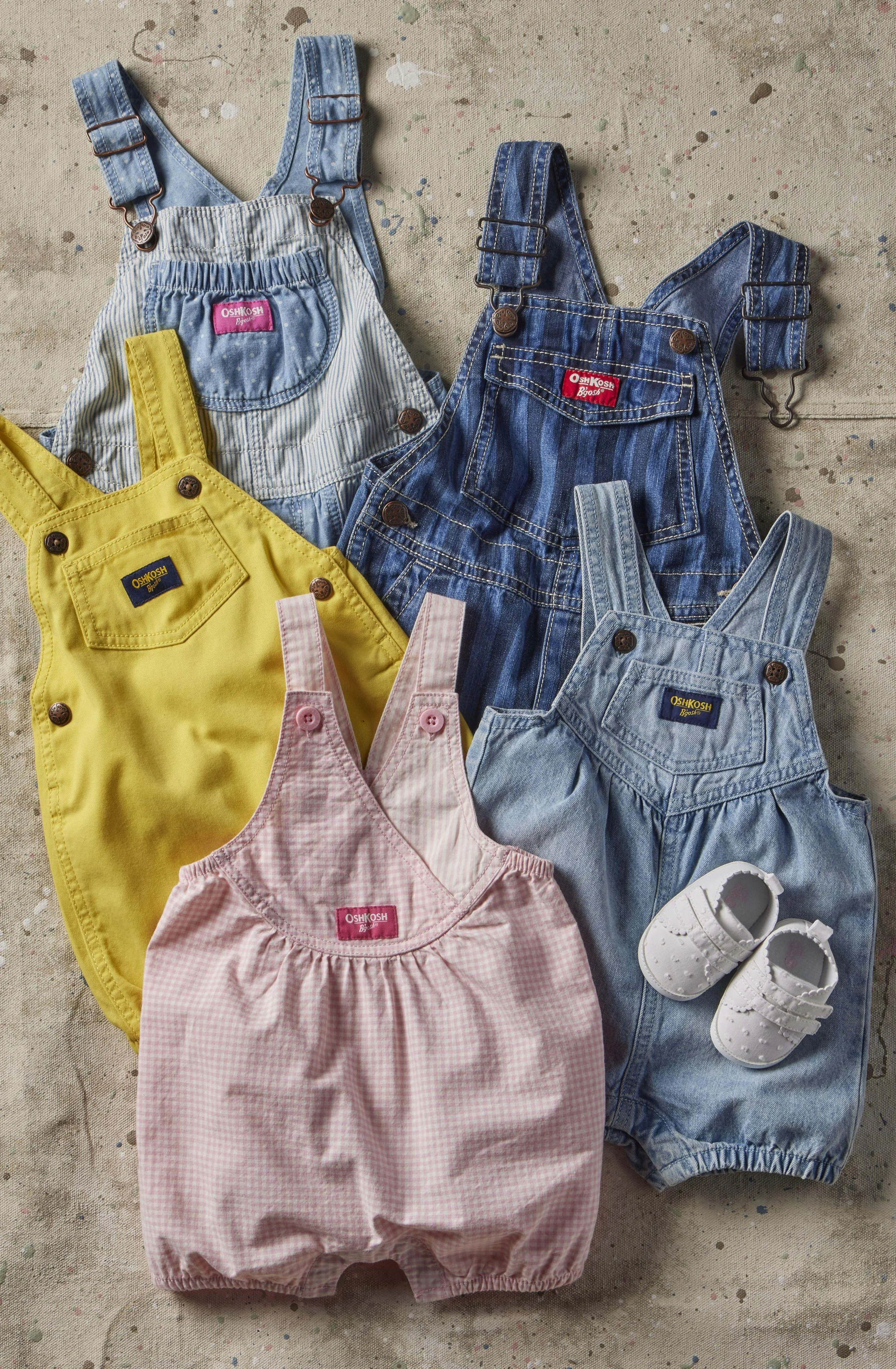 Five pairs of OshKosh overalls in different colors and patterns, arranged on a speckled concrete floor.