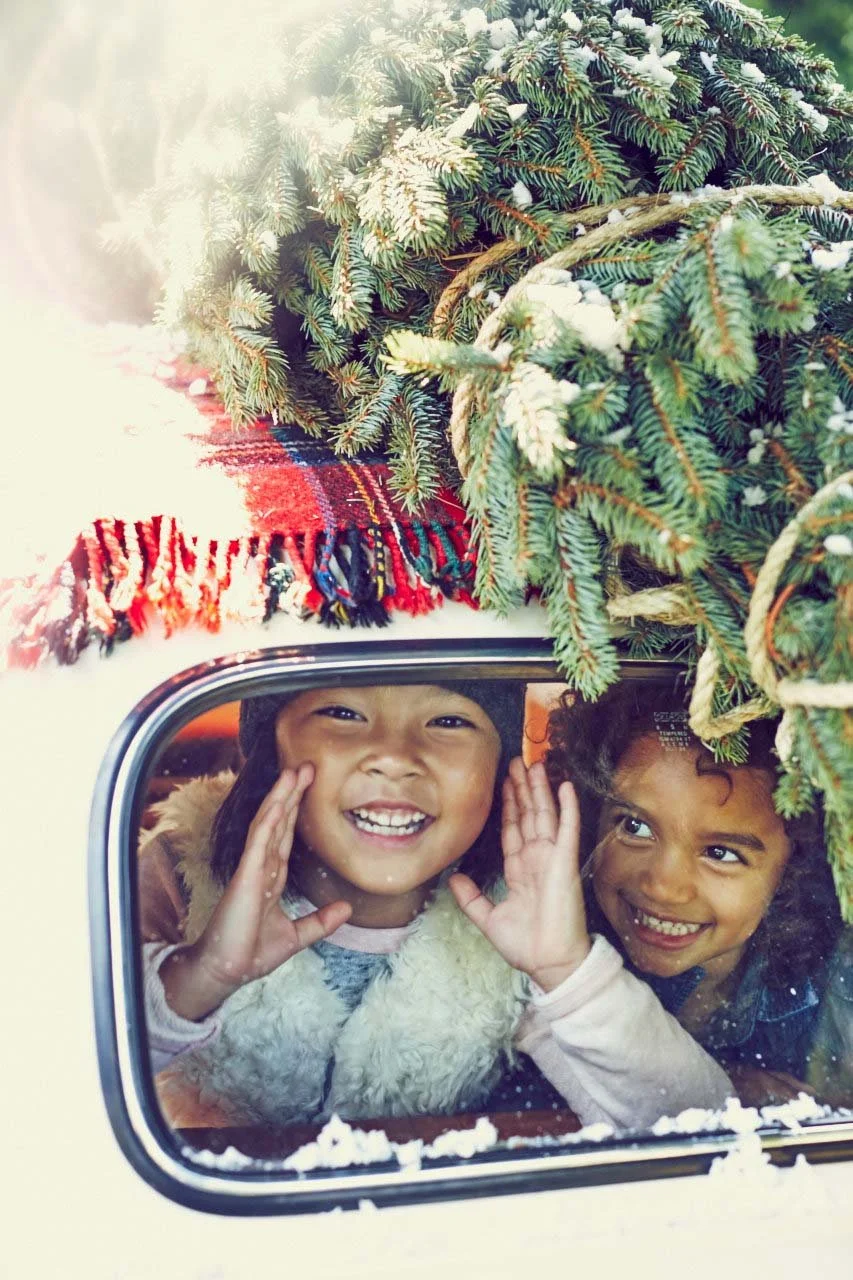 Two children looking through a window of a vehicle decorated with a Christmas tree and a red plaid blanket, smiling.
