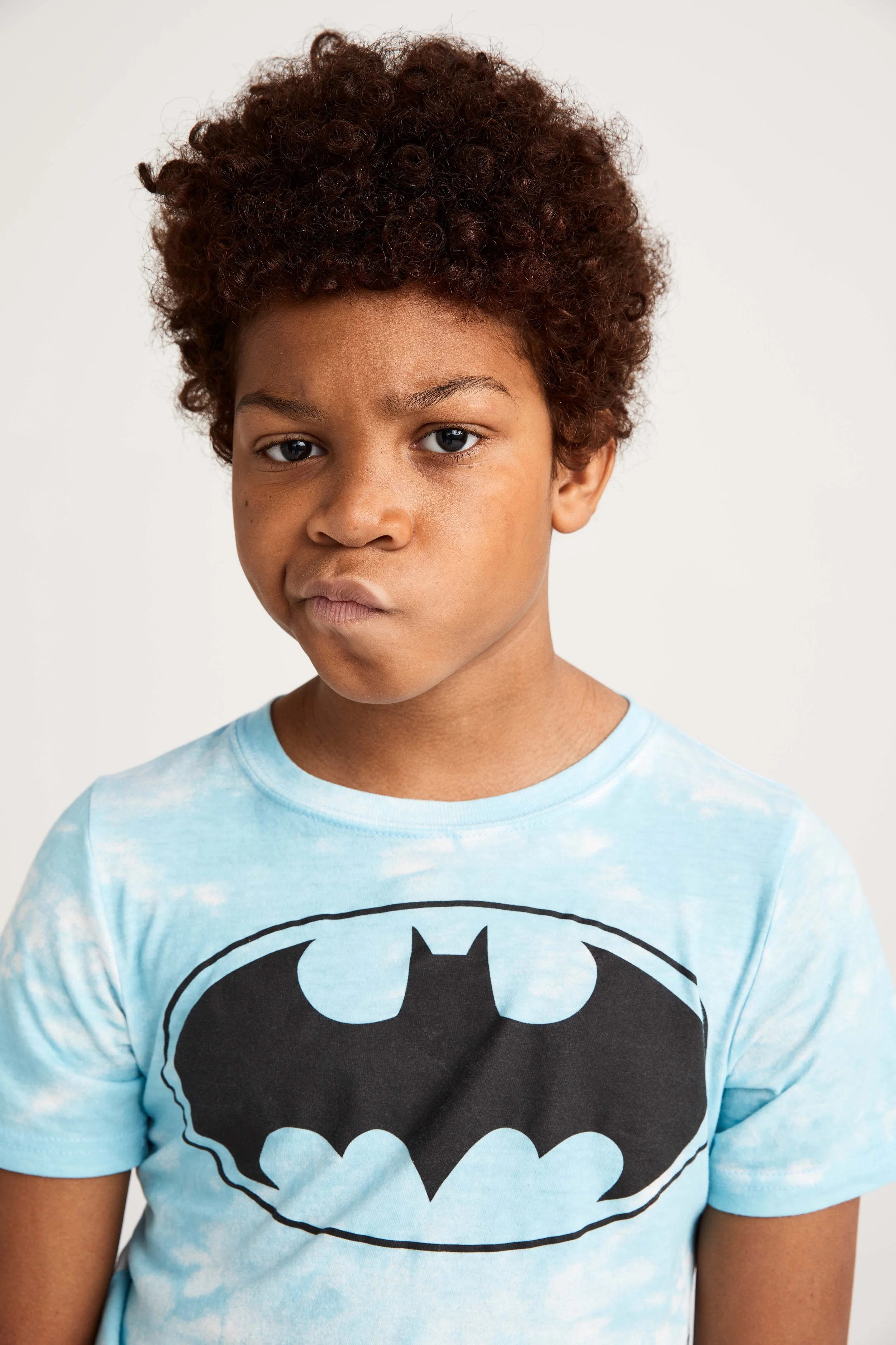 Close-up of a young boy with curly brown hair wearing a light blue Batman T-shirt, against a plain white background.
