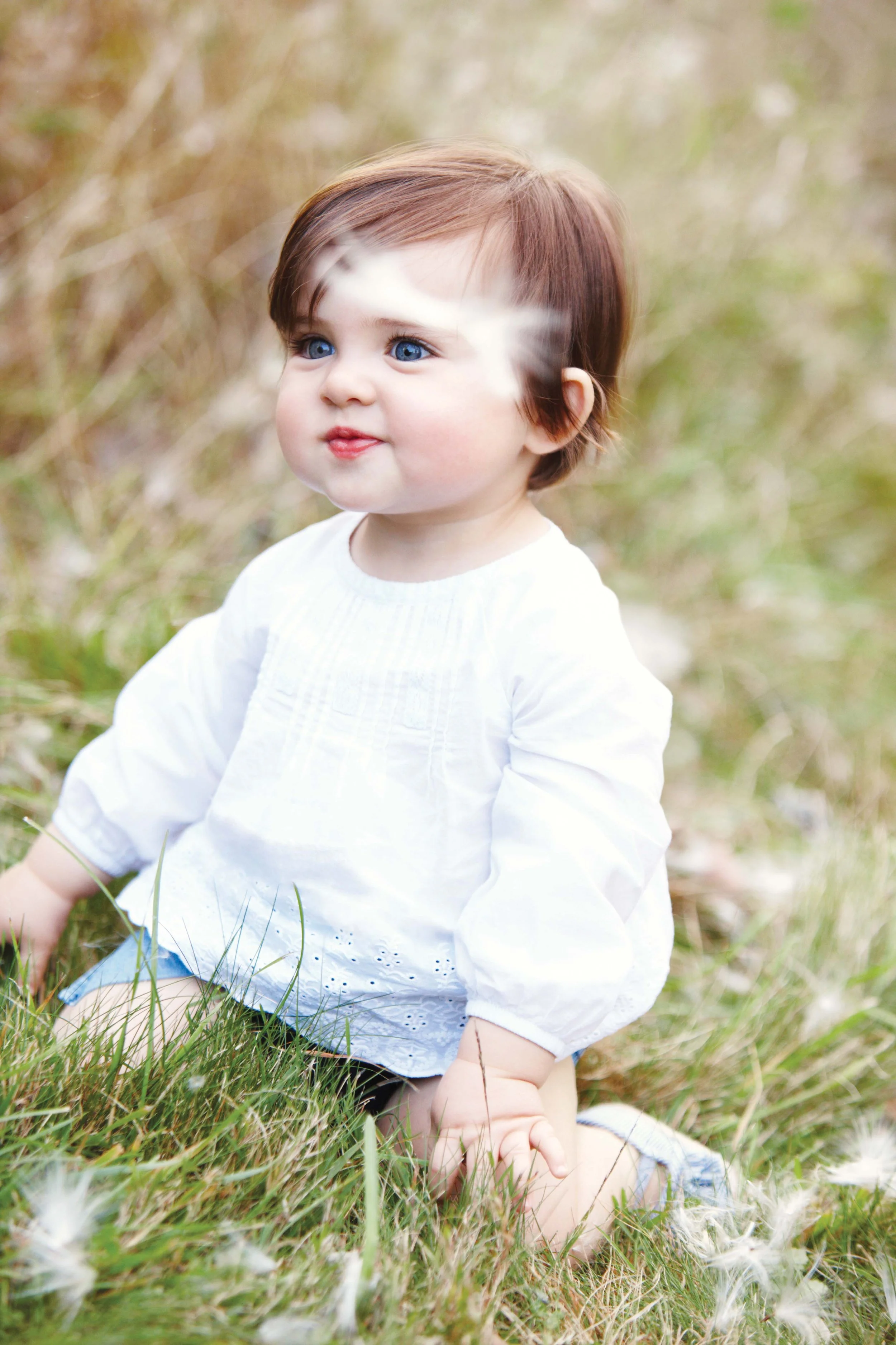 A young child with brown hair and bright blue eyes sitting on grass outdoors, wearing a white long sleeve top, with a blurred background of grass and foliage.