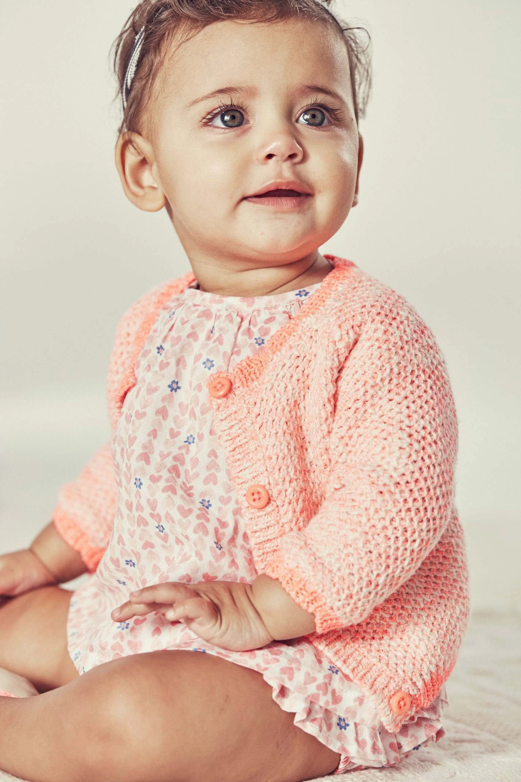 A young girl with curly hair wearing a pink knitted sweater and a floral dress, sitting on a person's lap with her hands resting on her knee.