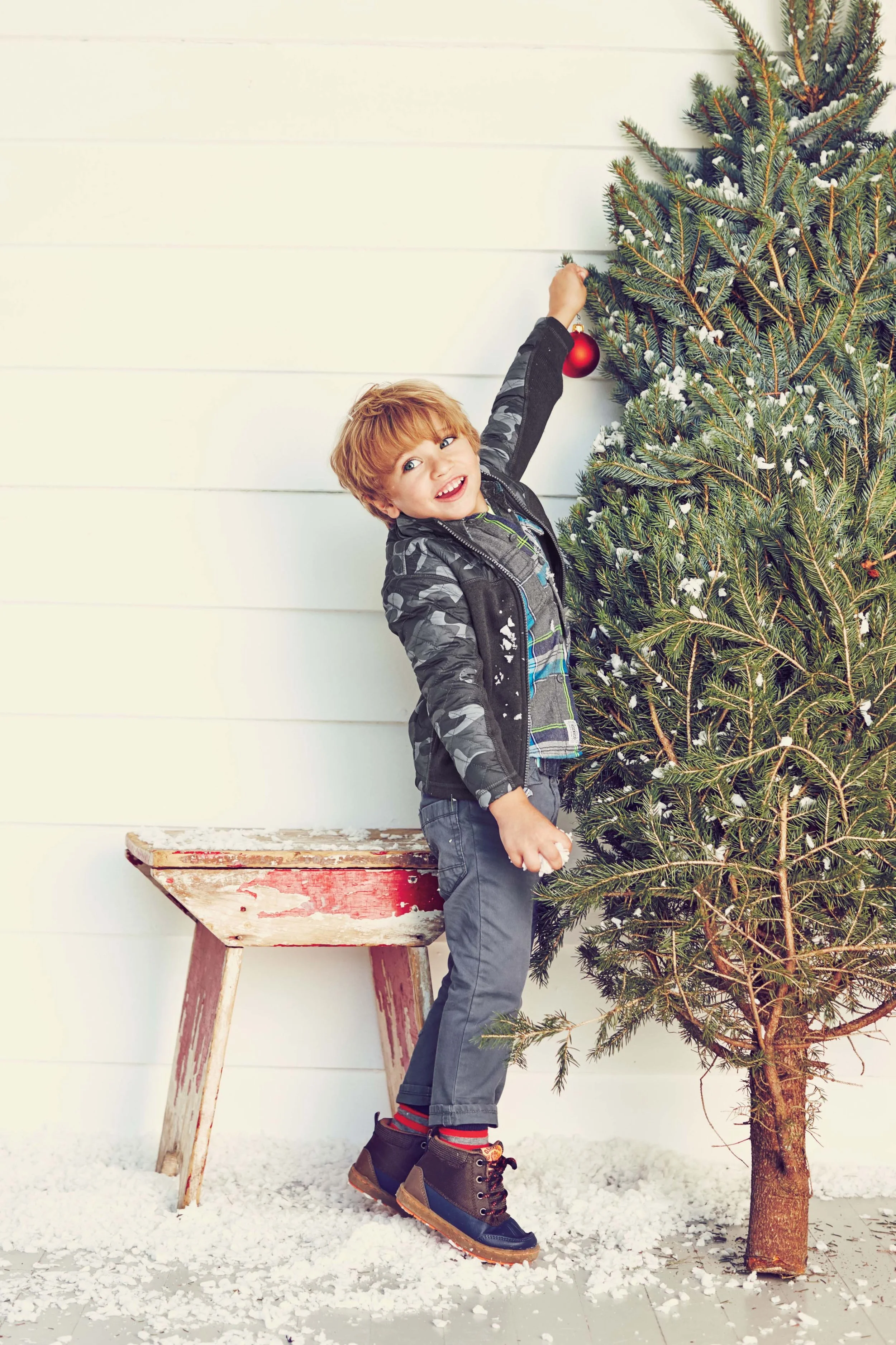 A young boy with red hair and a big smile decorating a Christmas tree with a red ornament, standing on the floor next to a rustic wooden stool, with fake snow on the ground and a white wooden wall background.
