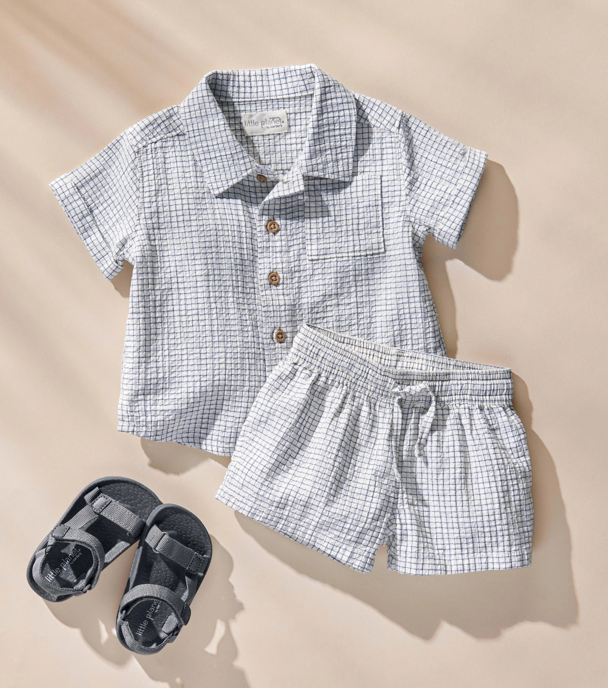 A children's outfit laid out on a beige surface, consisting of a light blue and white checkered short-sleeve button-up shirt, matching checkered shorts, and a pair of black and gray sandals with velcro straps.