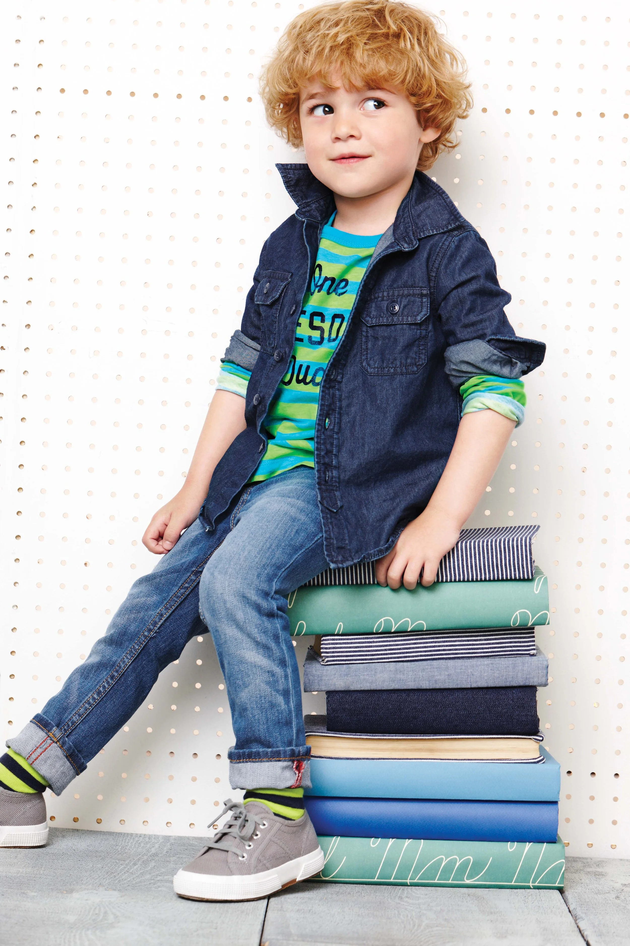 A young boy with curly red hair sitting on a stack of colorful storage boxes against a white pegboard background, wearing a denim jacket, striped shirt, jeans, and sneakers.