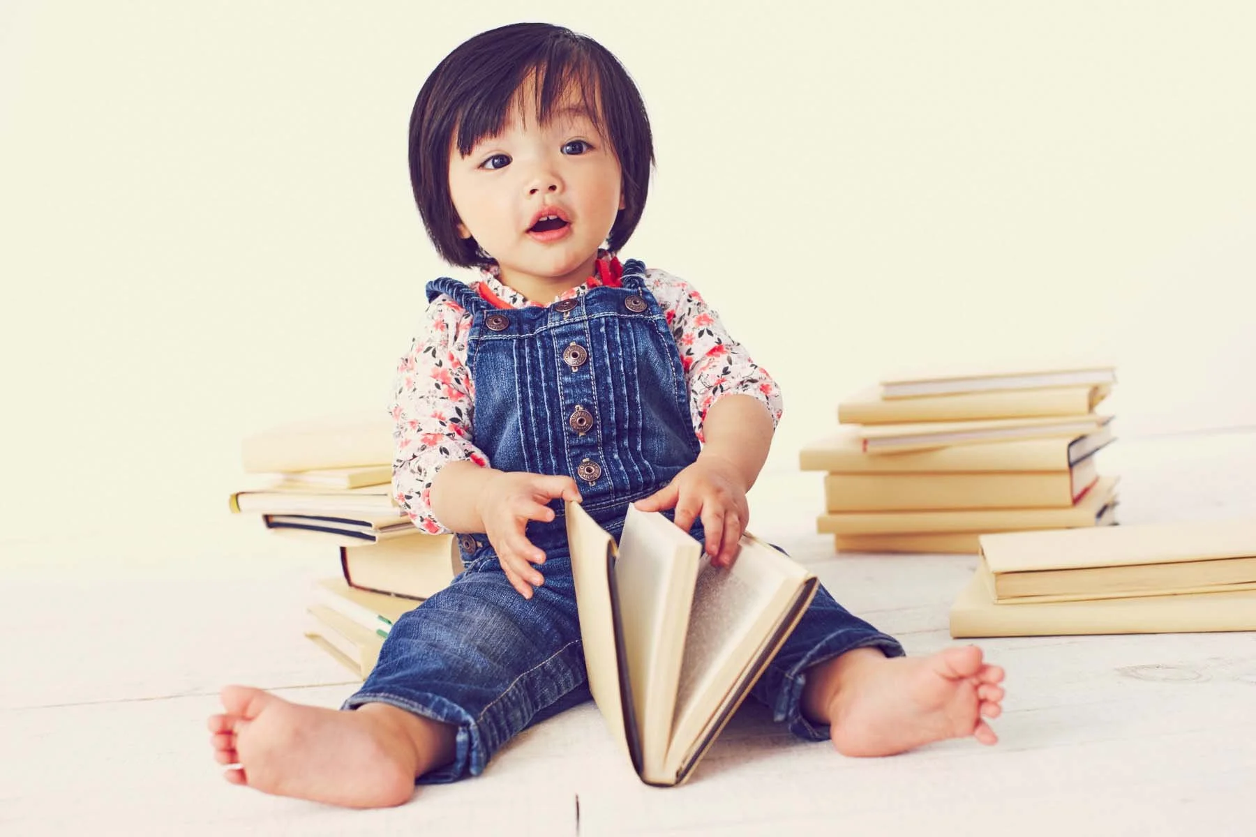 A young child with dark hair, wearing a floral shirt and denim overalls, sitting on a white floor, holding an open book and surrounded by stacked books, with a pale yellow background.