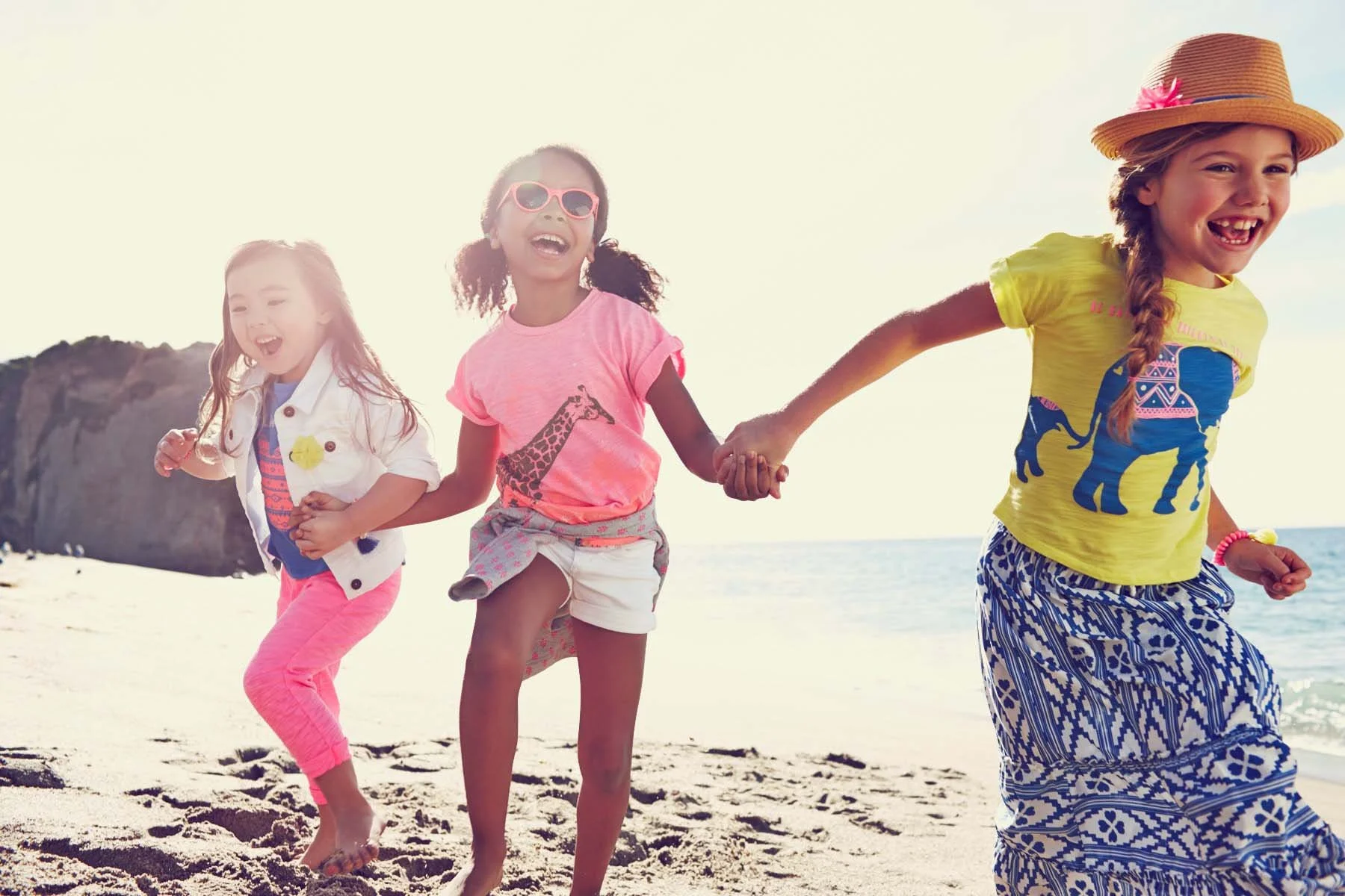 Three young girls playing and running hand-in-hand along the beach with a rocky cliff and ocean in the background, smiling and enjoying a sunny day.