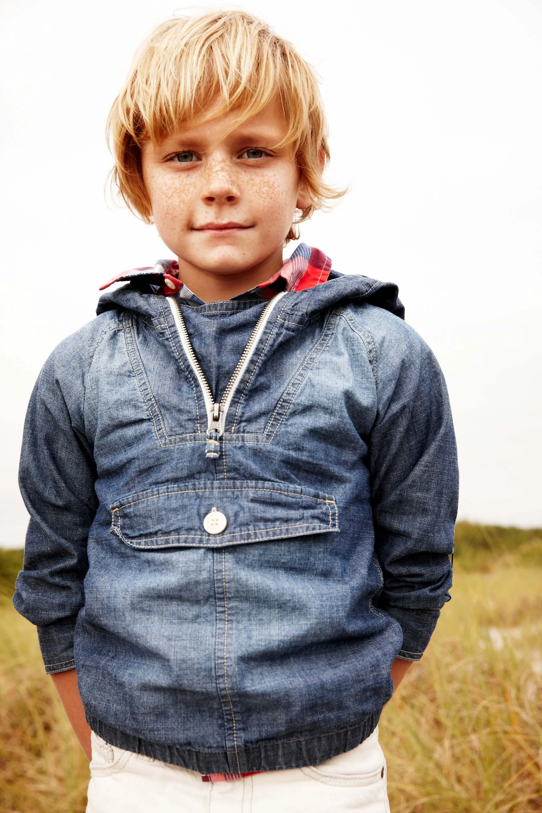 A young boy with blonde hair, freckles, wearing a denim jacket, standing outdoors in a field, with a cloudy sky in the background.