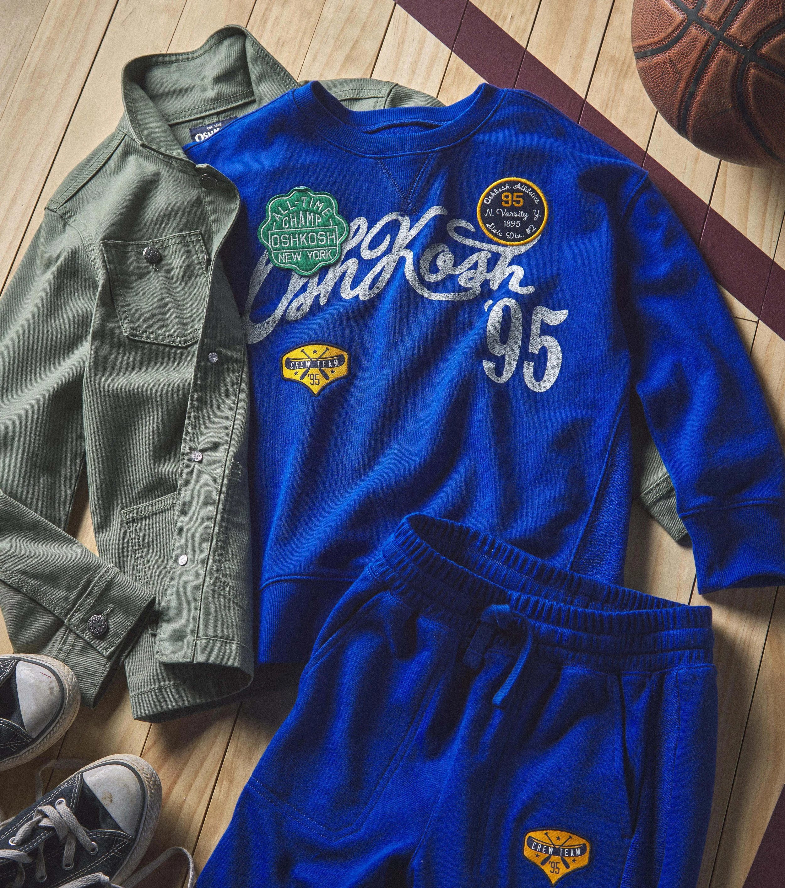 A green jacket and a blue sweatshirt with patches and embroidery are lying on a wooden floor next to a basketball and a pair of sneakers.