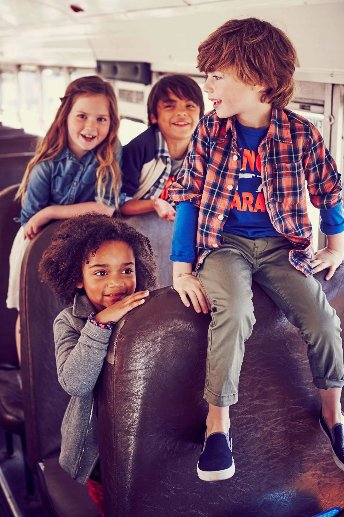 Four children on a school bus, with one girl sitting on the armrest of a bus seat and three children behind her leaning on the seat back, smiling and looking at each other.