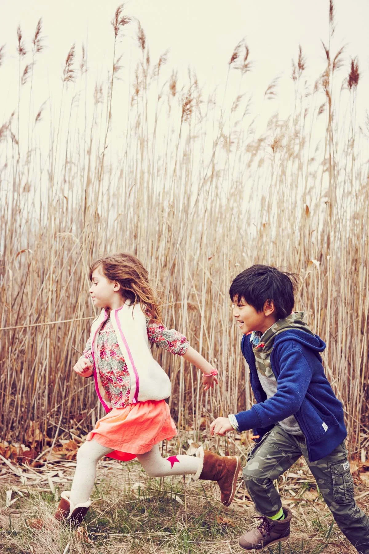 Two children, a girl and a boy, running and playing through a field of tall, dry grass, holding hands and smiling during daytime.