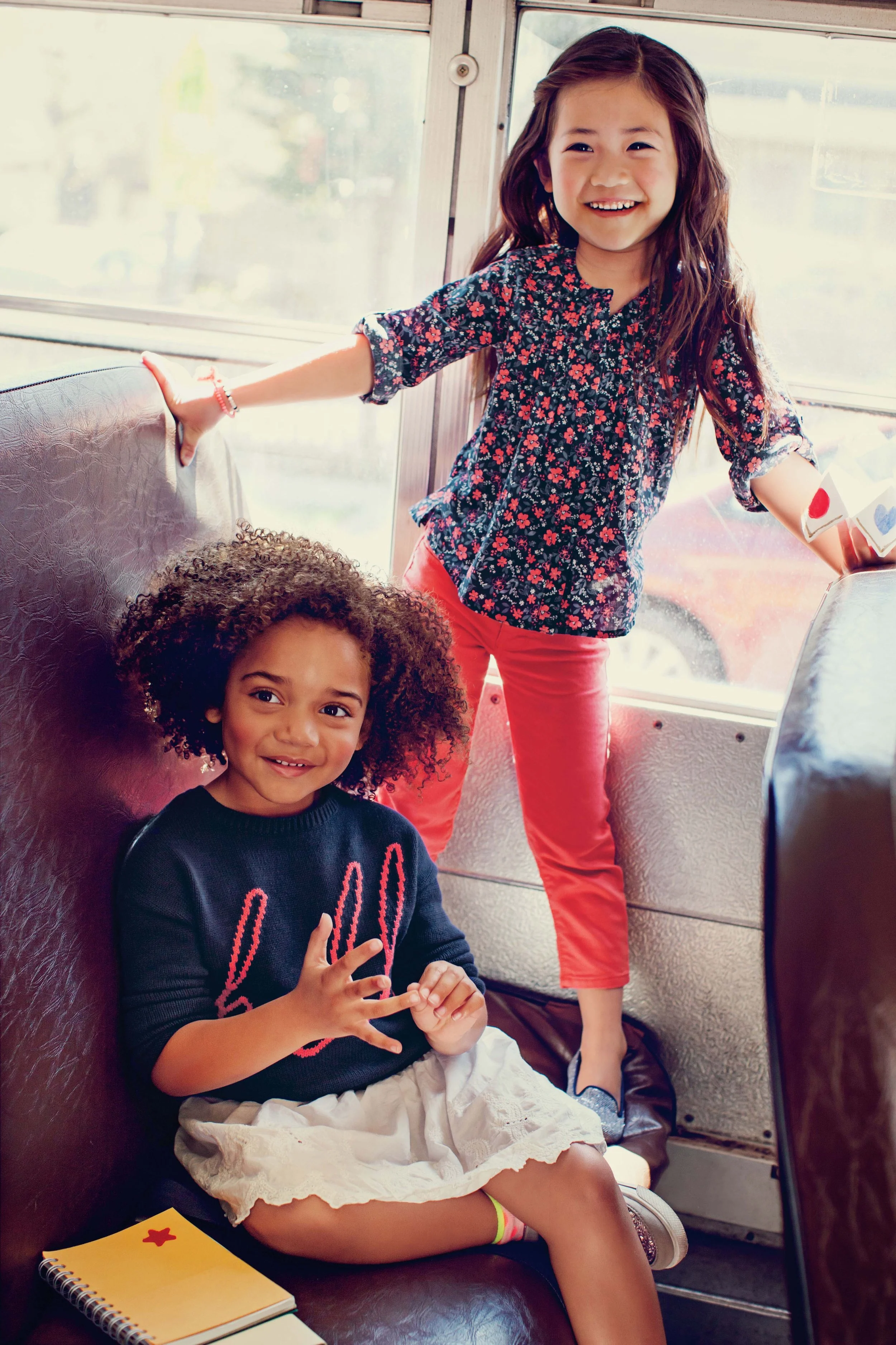 A young girl and a young boy sitting on a bus. The girl is smiling and standing, holding onto the bus's window frame, wearing a floral shirt and pink pants. The boy is sitting, making a heart shape with his hands, wearing a dark sweater with pink wri