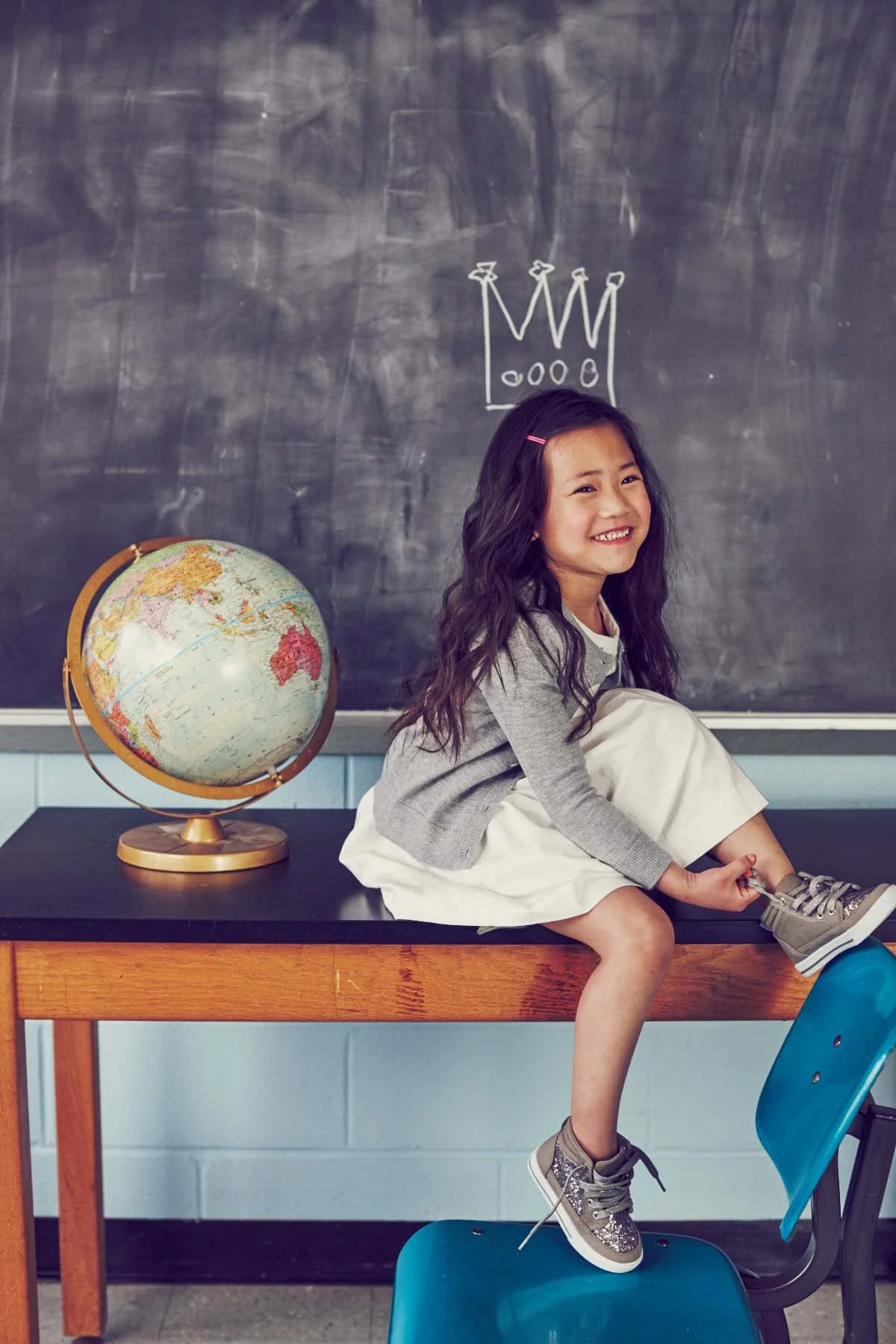 A smiling young girl with long dark hair sitting on a classroom desk, tying her shoelace. There is a globe and a chalkboard with a simple crown drawing behind her.