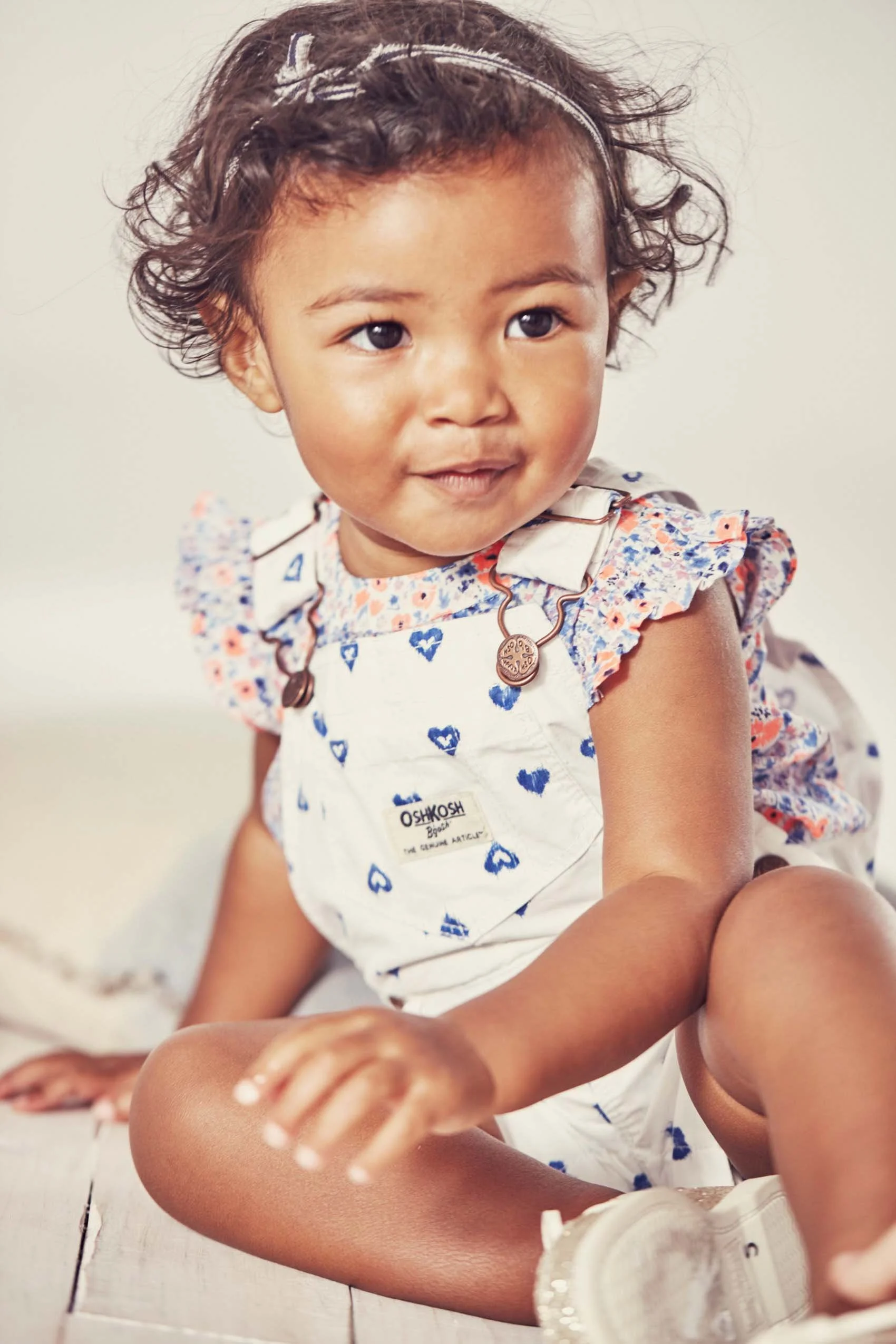 A young girl with curly brown hair wearing a headband, a white dress with blue and orange patterns, and a necklace sitting on a white wooden floor along with a container of ice cream.