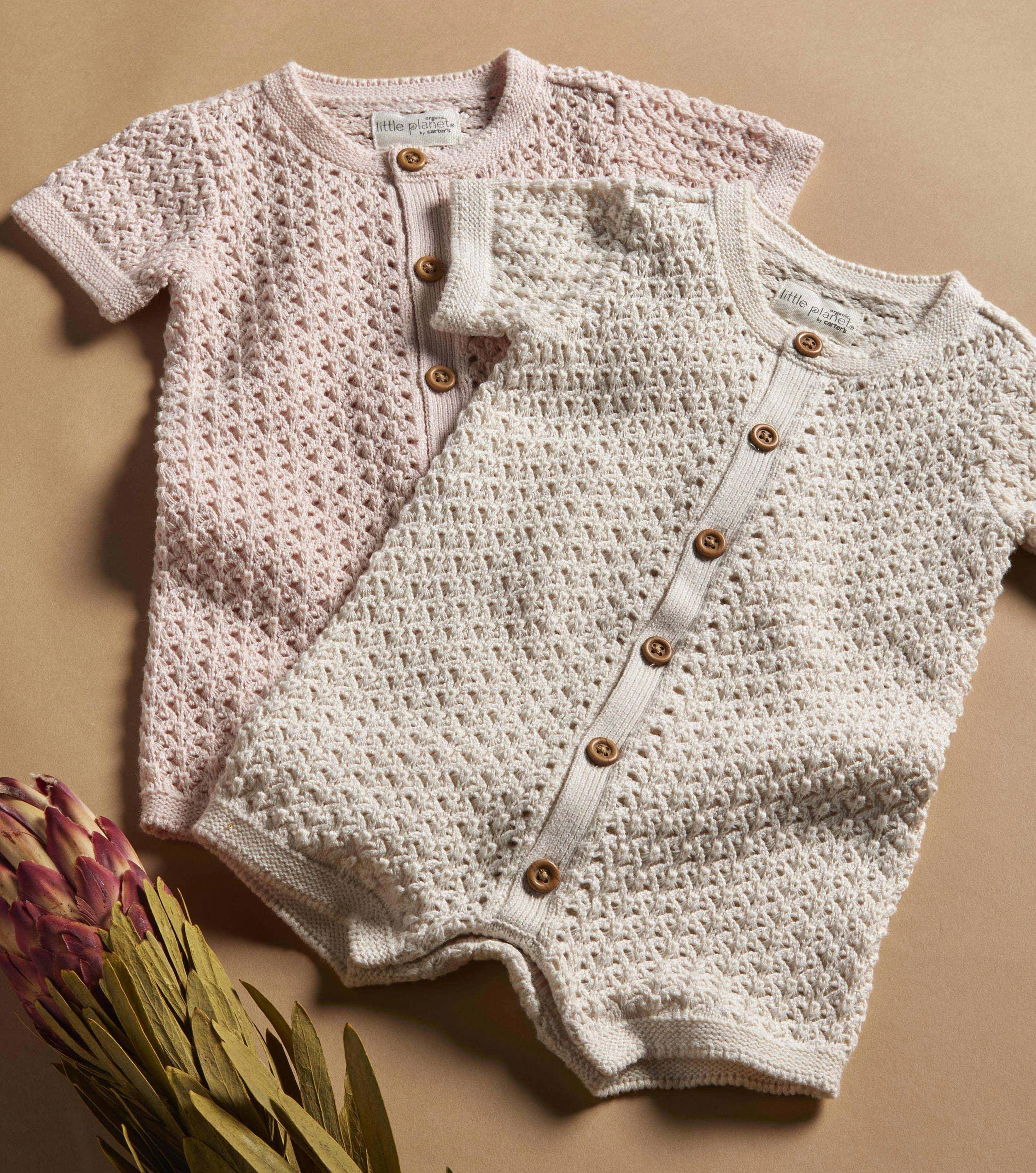 Two knitted baby cardigans, one pink and one cream, with buttons, laid out on a beige surface near dried flowers.