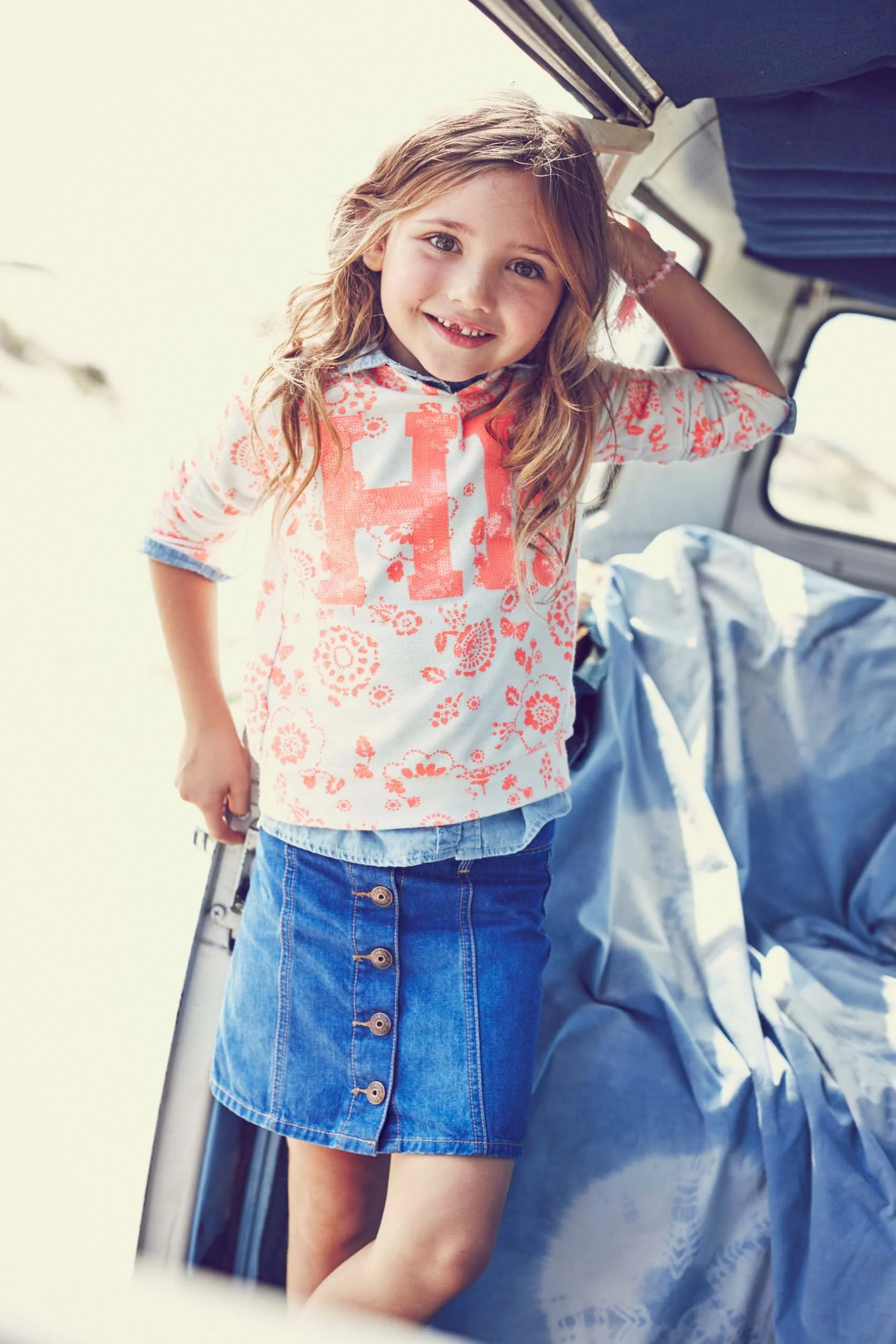 A young girl with long wavy hair and a floral top stands in a room with a window, smiling at the camera, as she leans against a door frame near a bed with rumpled bedding.