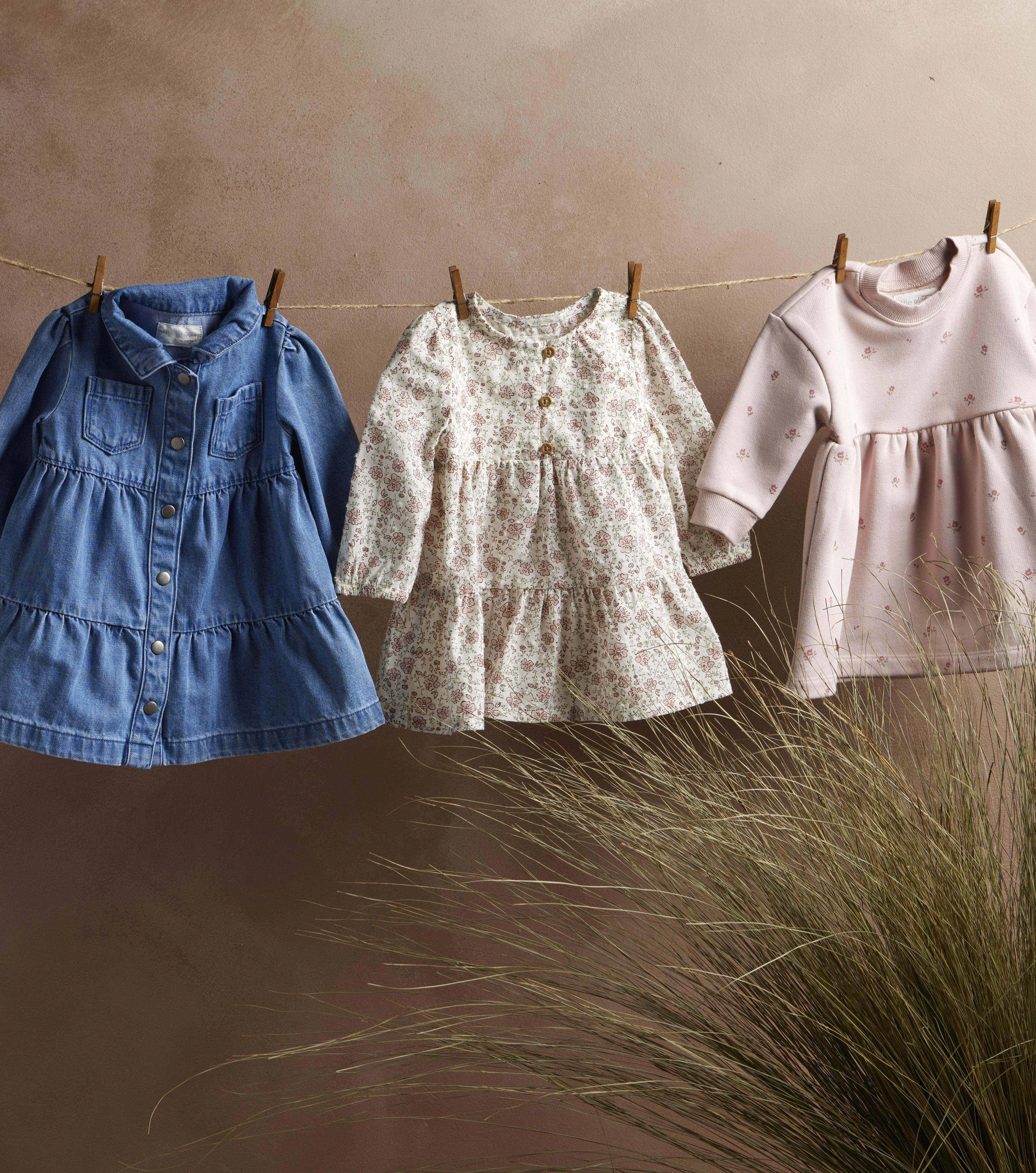 Three children's dresses hanging on a string with clothespins against a beige wall, with grass in the foreground.