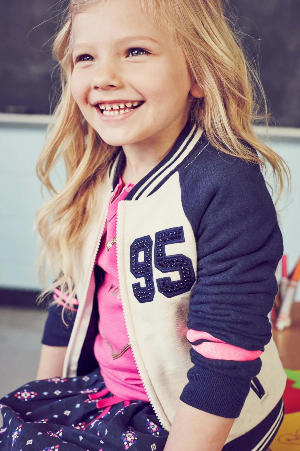 Smiling young girl with light skin and long red hair, wearing a baseball jacket with the number 95, pink shirt, and patterned pants, sitting in a classroom.