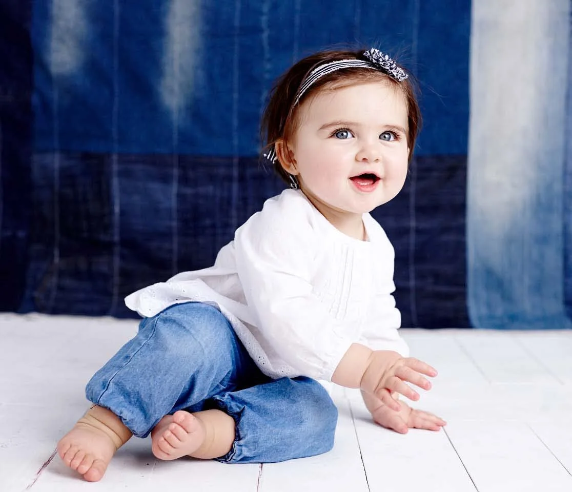 A smiling toddler girl with blue eyes, wearing a white top, denim pants, and a headband, sitting on the white wooden floor in front of a blue and beige fabric backdrop.