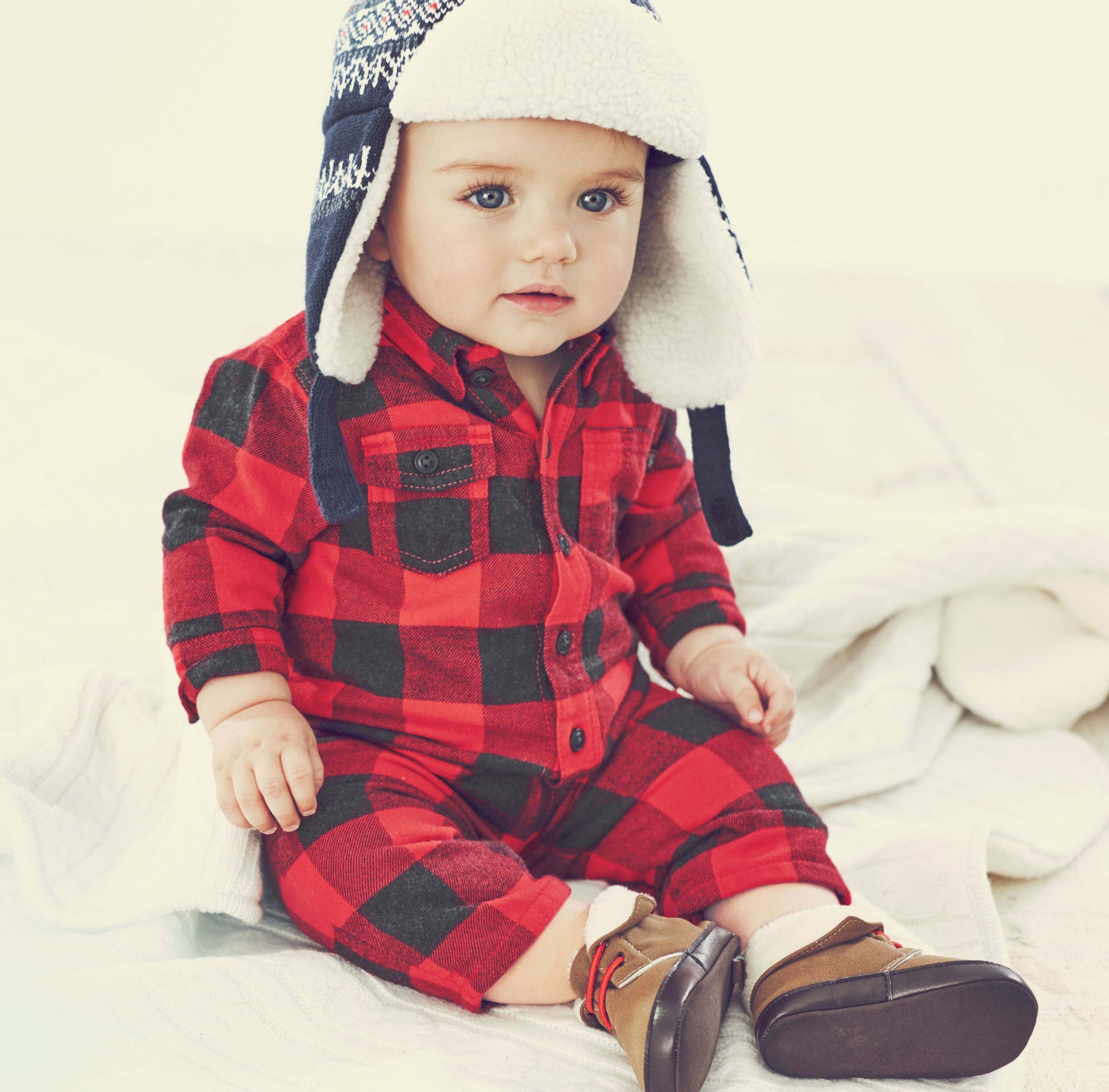 A young child with blue eyes wearing a black, red, and gray plaid shirt, brown shoes, and a white and navy knit hat, sitting on a white surface.