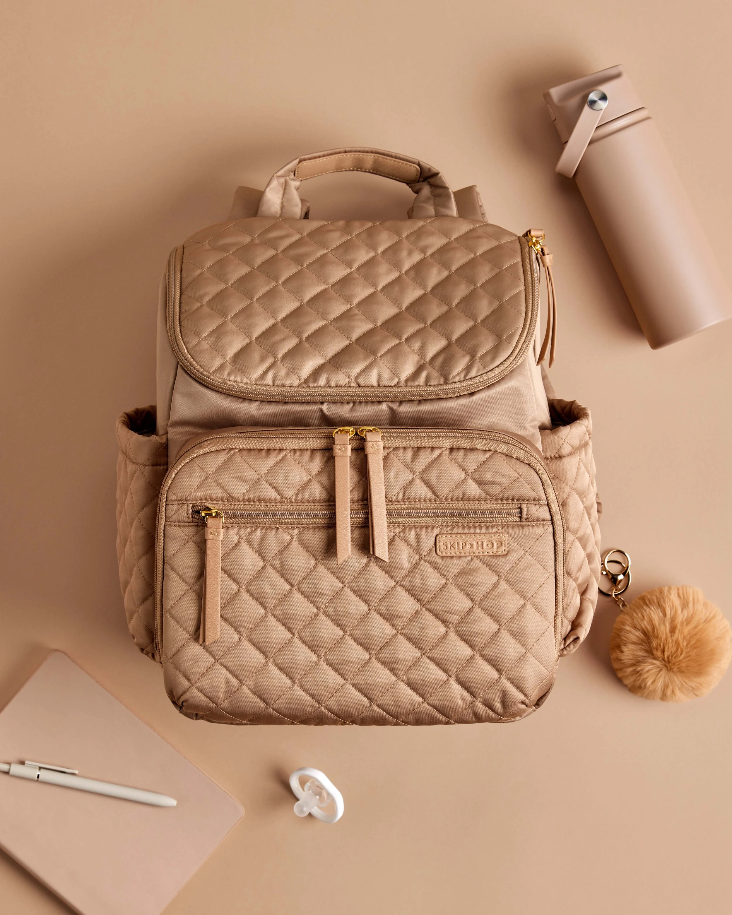 A beige quilted backpack with a handle and zipper pockets, a beige water bottle with a strap, a white pen, a pair of earbuds, a closed laptop, and a fluffy pom-pom accessory on a beige surface.